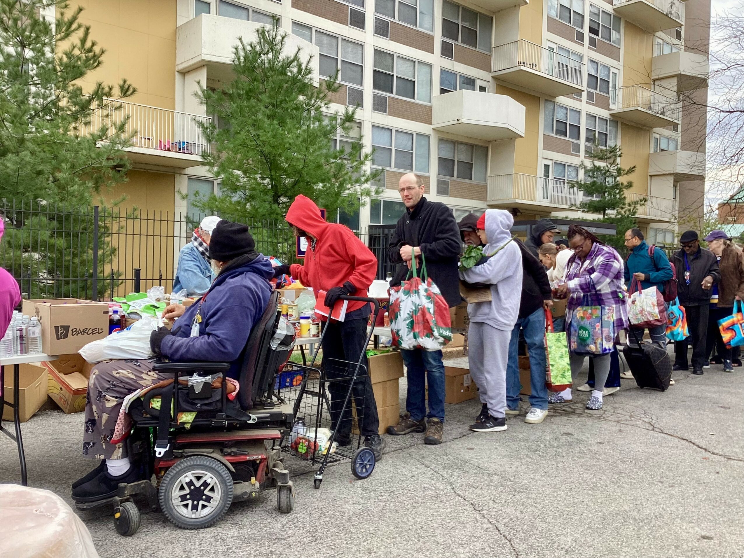 People line up at an outdoor market with tables and boxes, in front of a multi-story apartment building, some in winter clothing.