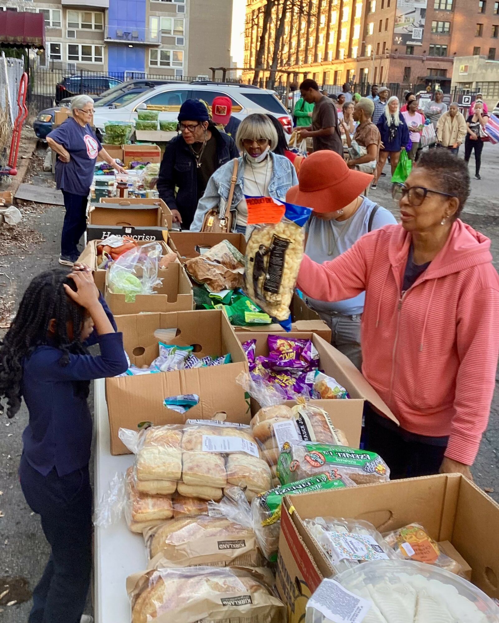 People shopping at an outdoor market with tables of bread, snacks, and groceries, in an urban setting with buildings and cars.