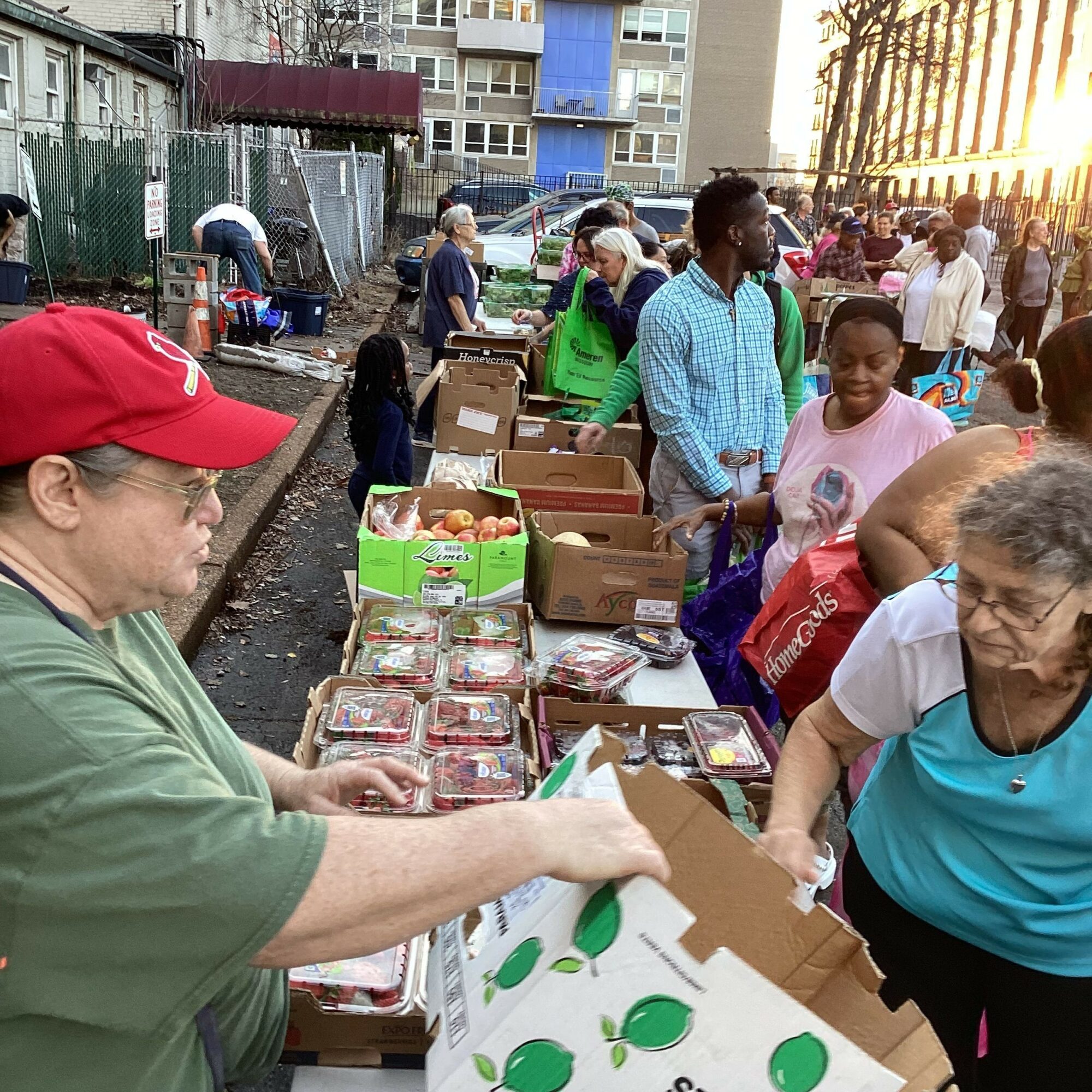 People gather around tables with boxes and items outdoors, with buildings and sunlight in the background.
