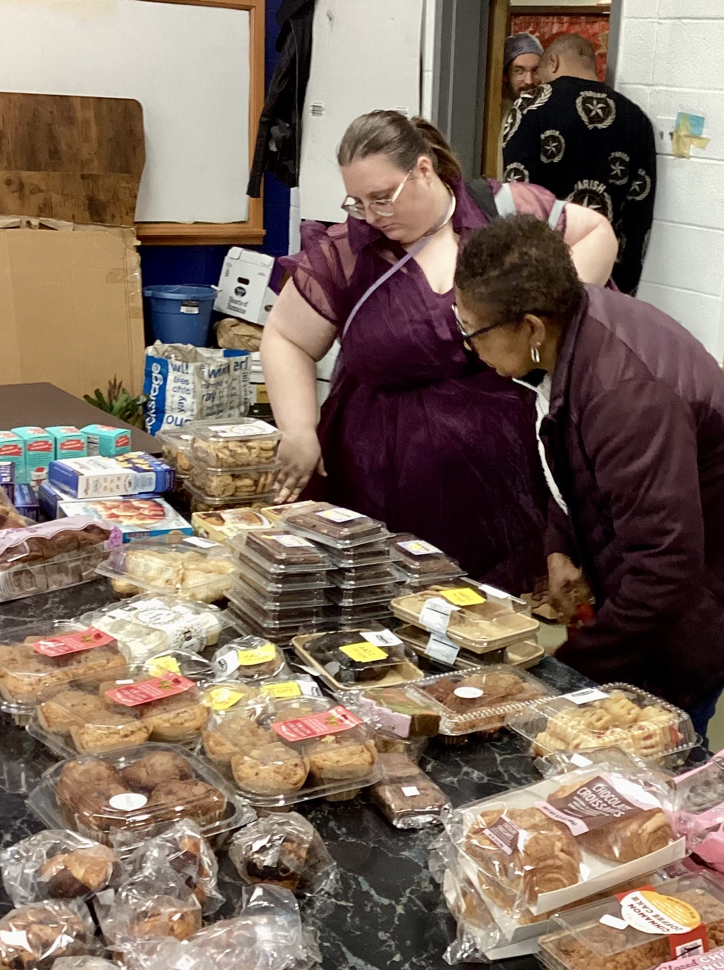 Two women look at baked goods and snacks on a table, with a person in the background near a door.