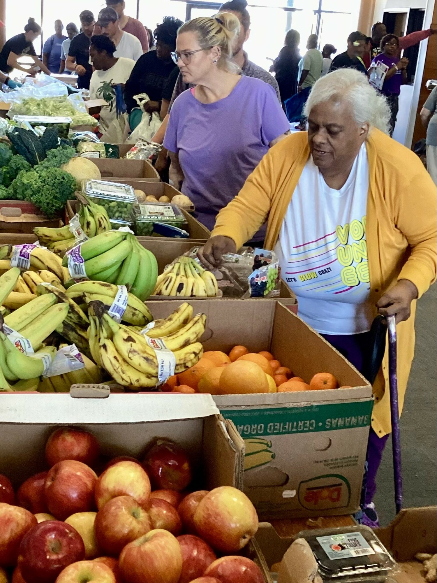 Two women shopping for fruits at a market stall with bananas, oranges, and apples, surrounded by other shoppers and produce displays.