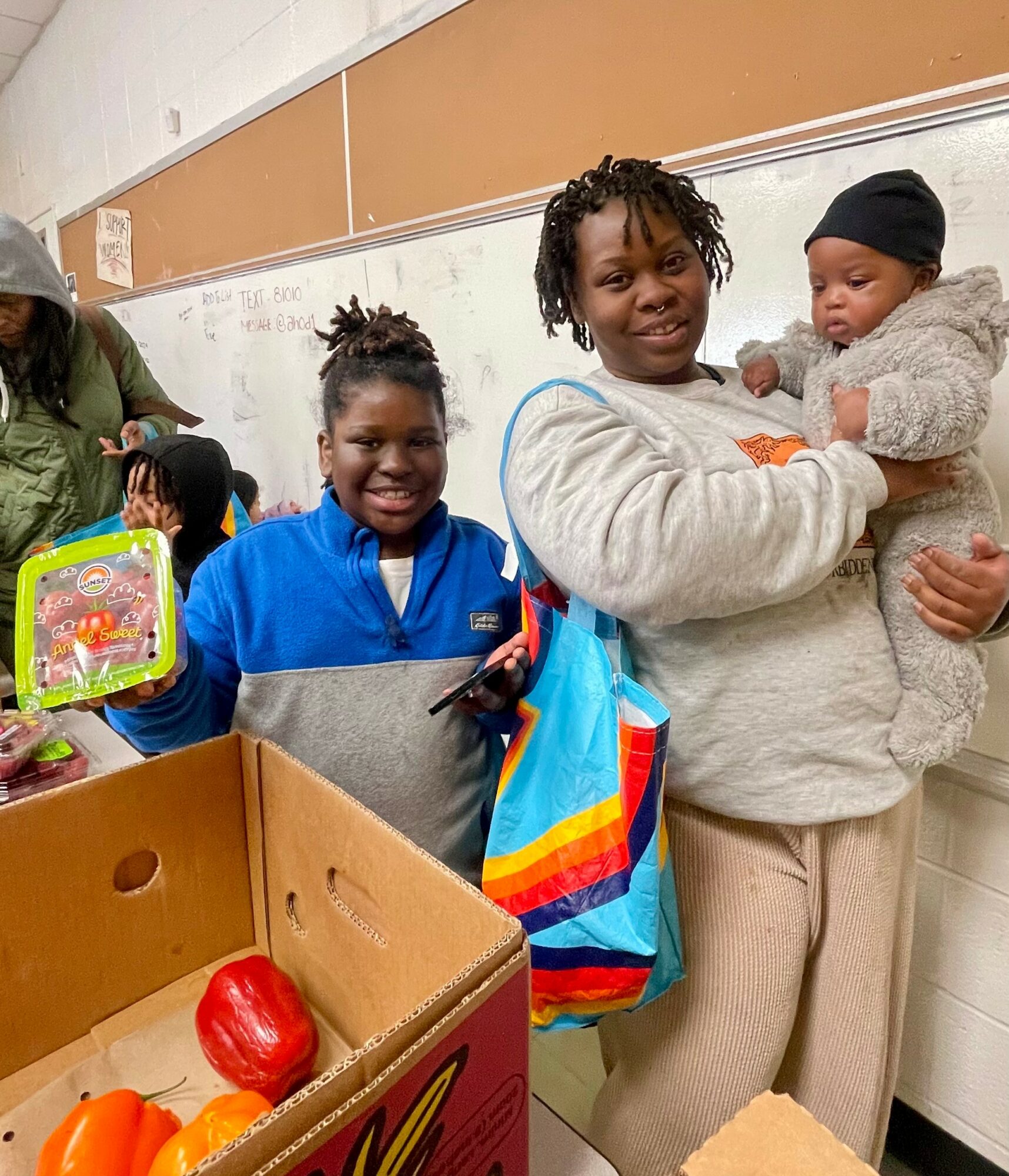 Two women and a child in a classroom, one woman holding a baby, smiling, with a box of toys and a colorful bag.