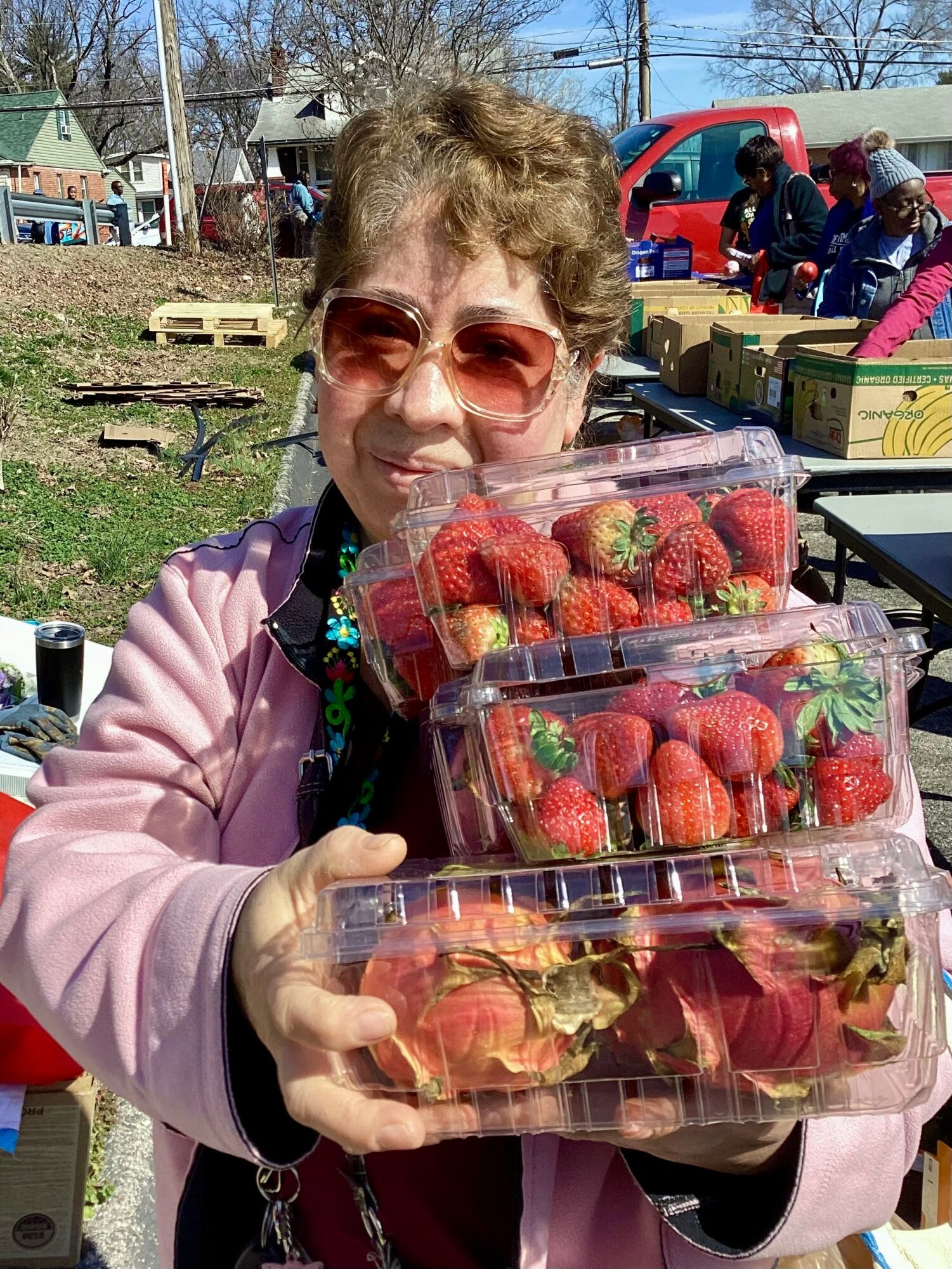 Woman holding three containers of strawberries outdoors, with people and a red vehicle in the background.