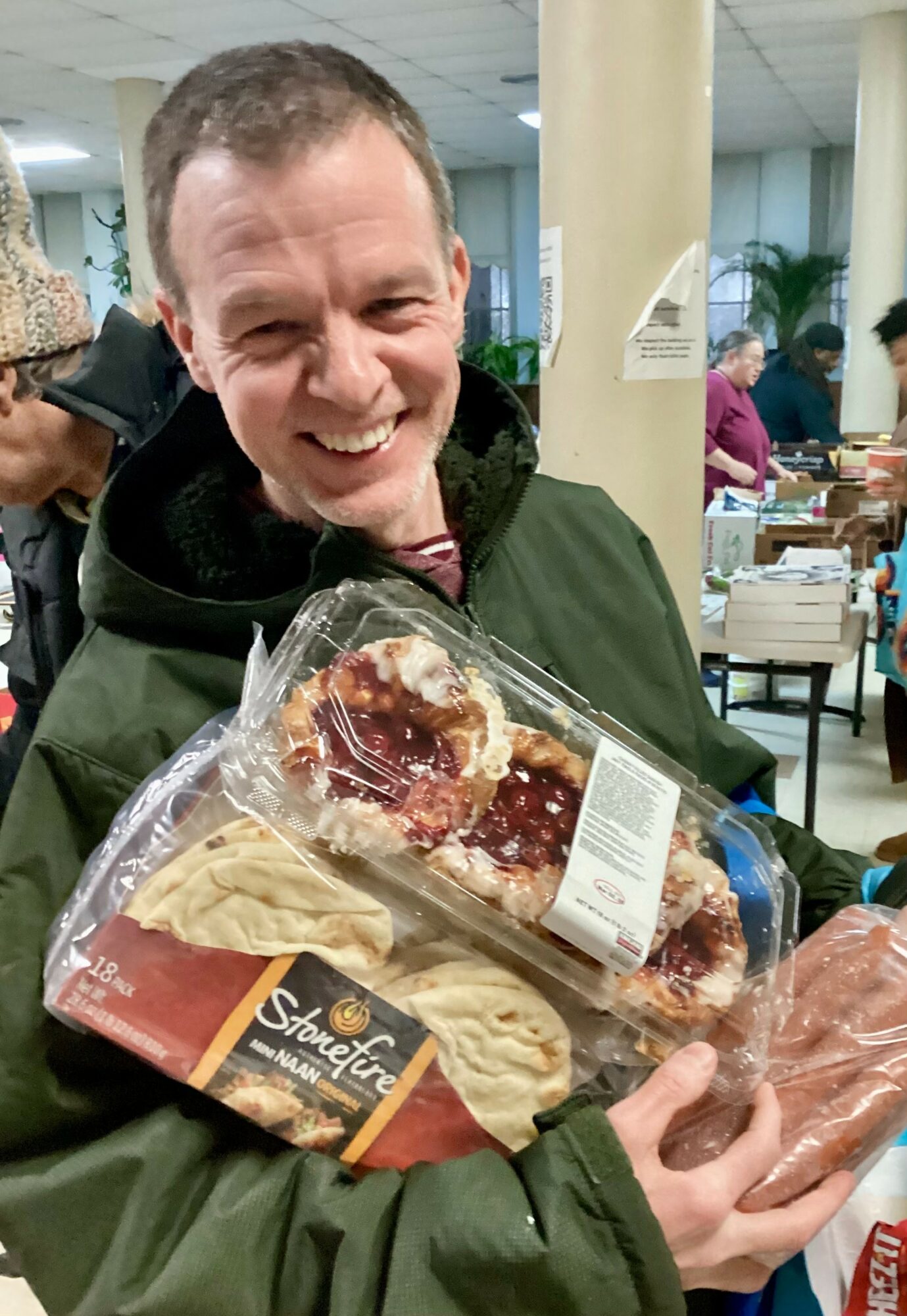 Smiling man holding packaged food items in a store or market, with other shoppers and shelves in the background.
