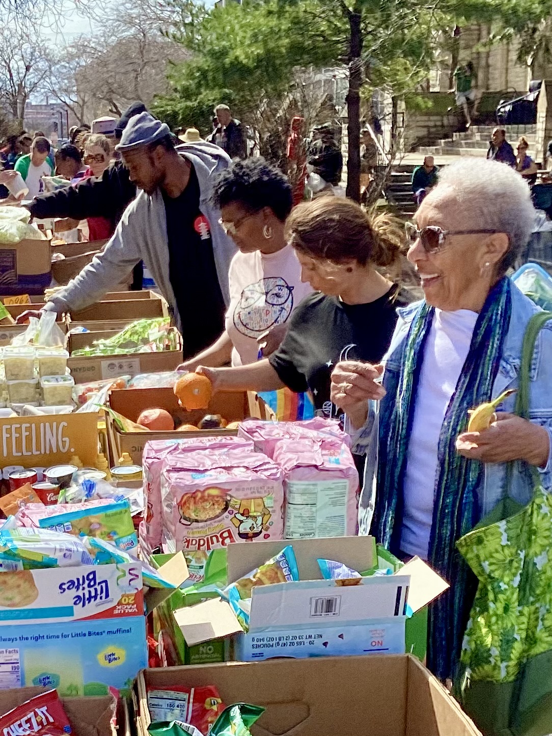 People shopping outdoors at a market with tables filled with food and goods, trees and buildings in background.