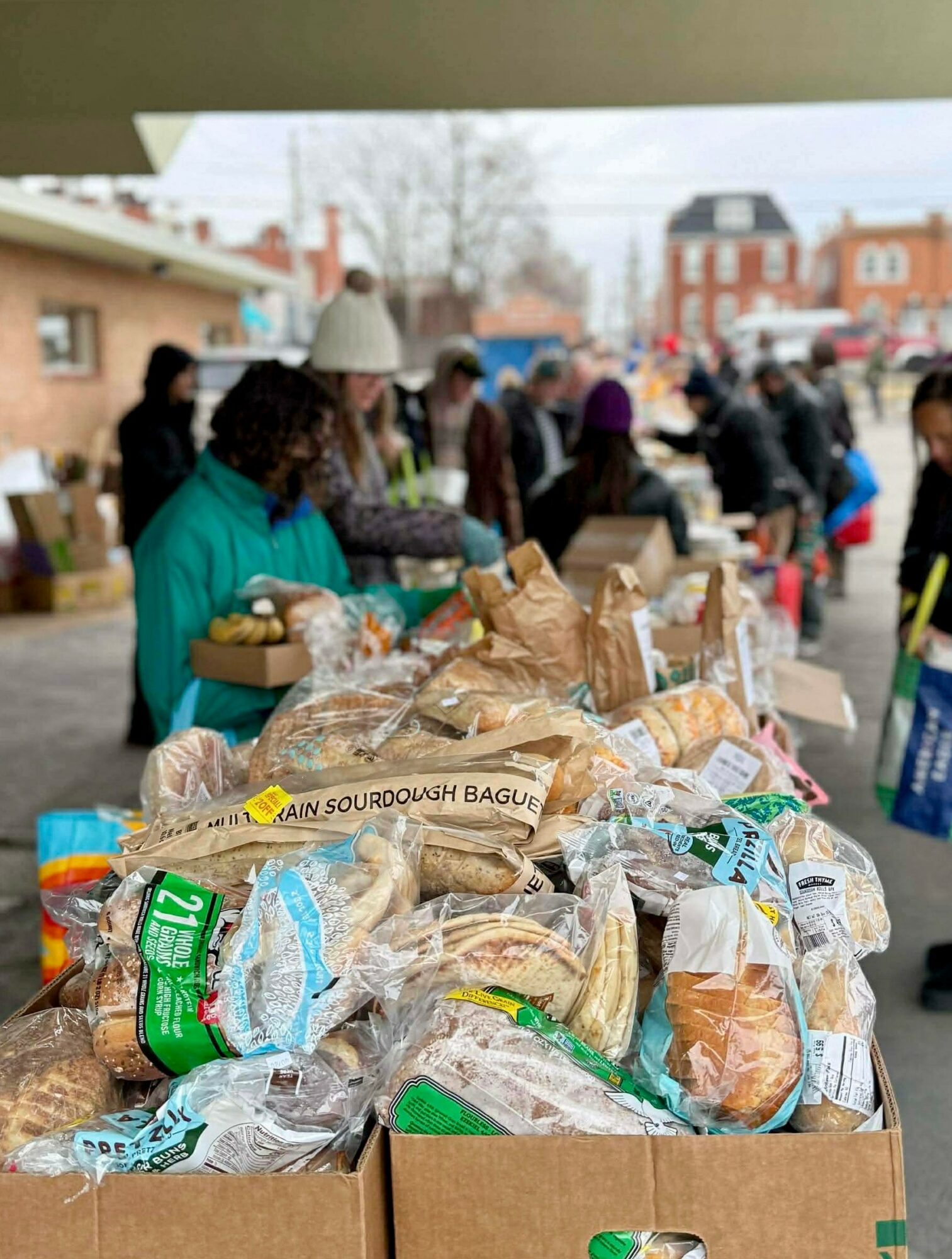 People line up at a table with bread and baked goods outdoors, some wearing hats and jackets, in a town setting.