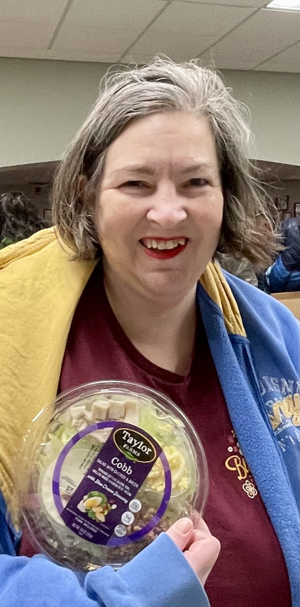Smiling woman with gray hair holding a container of food, wearing a yellow and blue jacket, indoors.