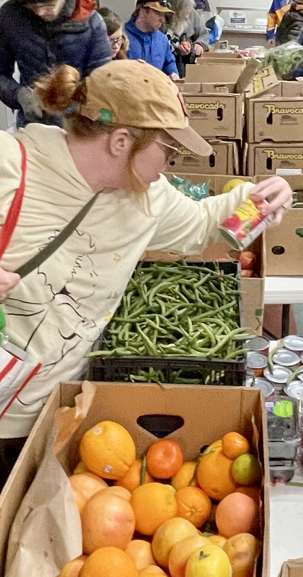 Person shopping for vegetables and fruits at a market stall, with boxes of oranges, apples, and green beans.