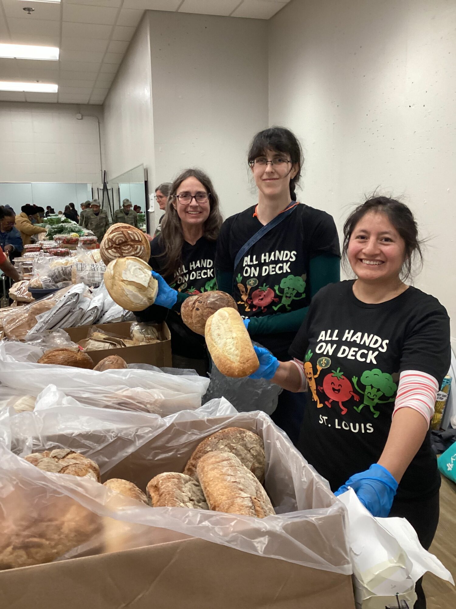 Three women stand behind a table with bread and baked goods, smiling at the camera, in a room with a white wall.