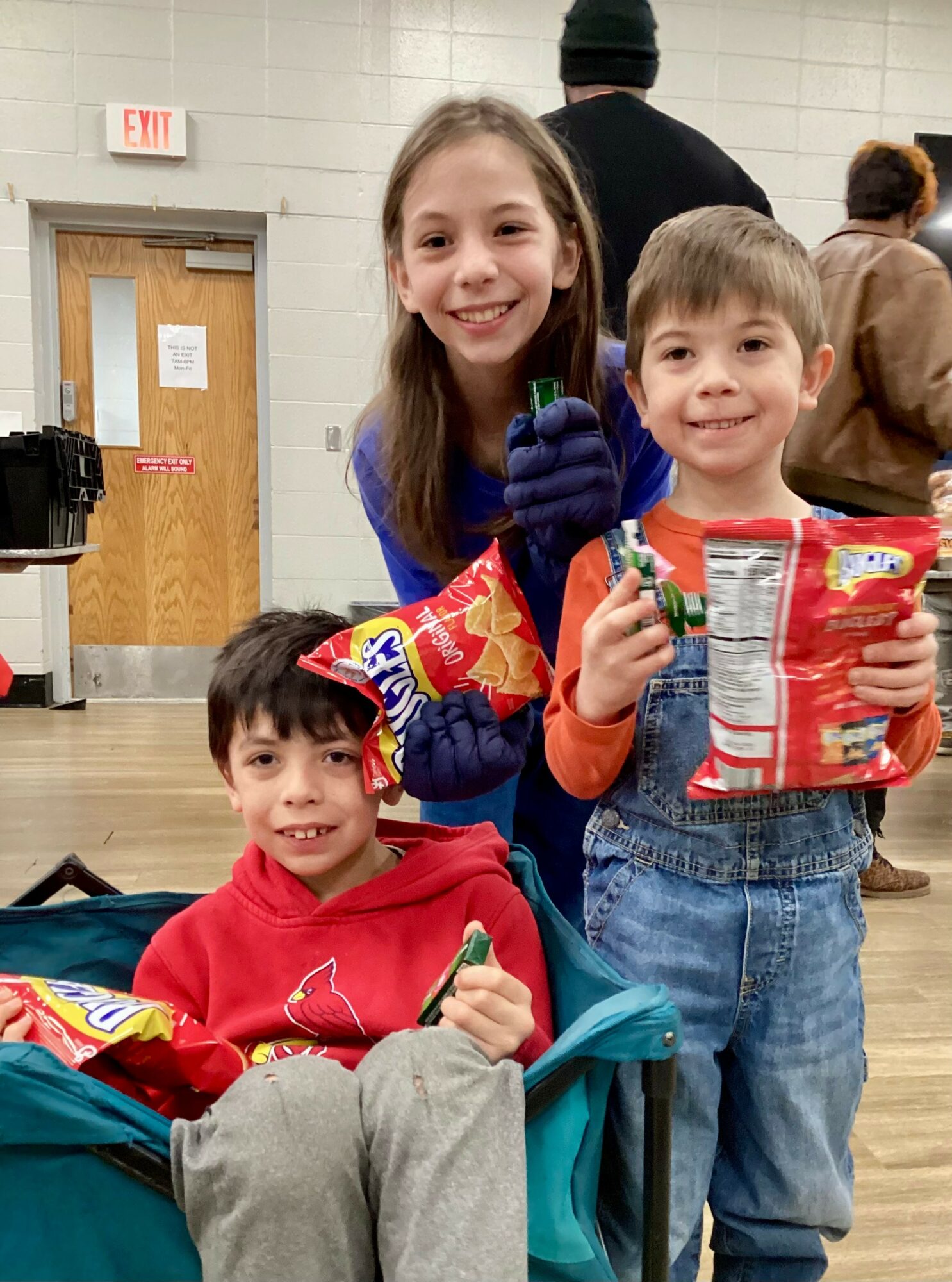 Three children smiling indoors, holding snacks and a drink, with a door and people in the background.