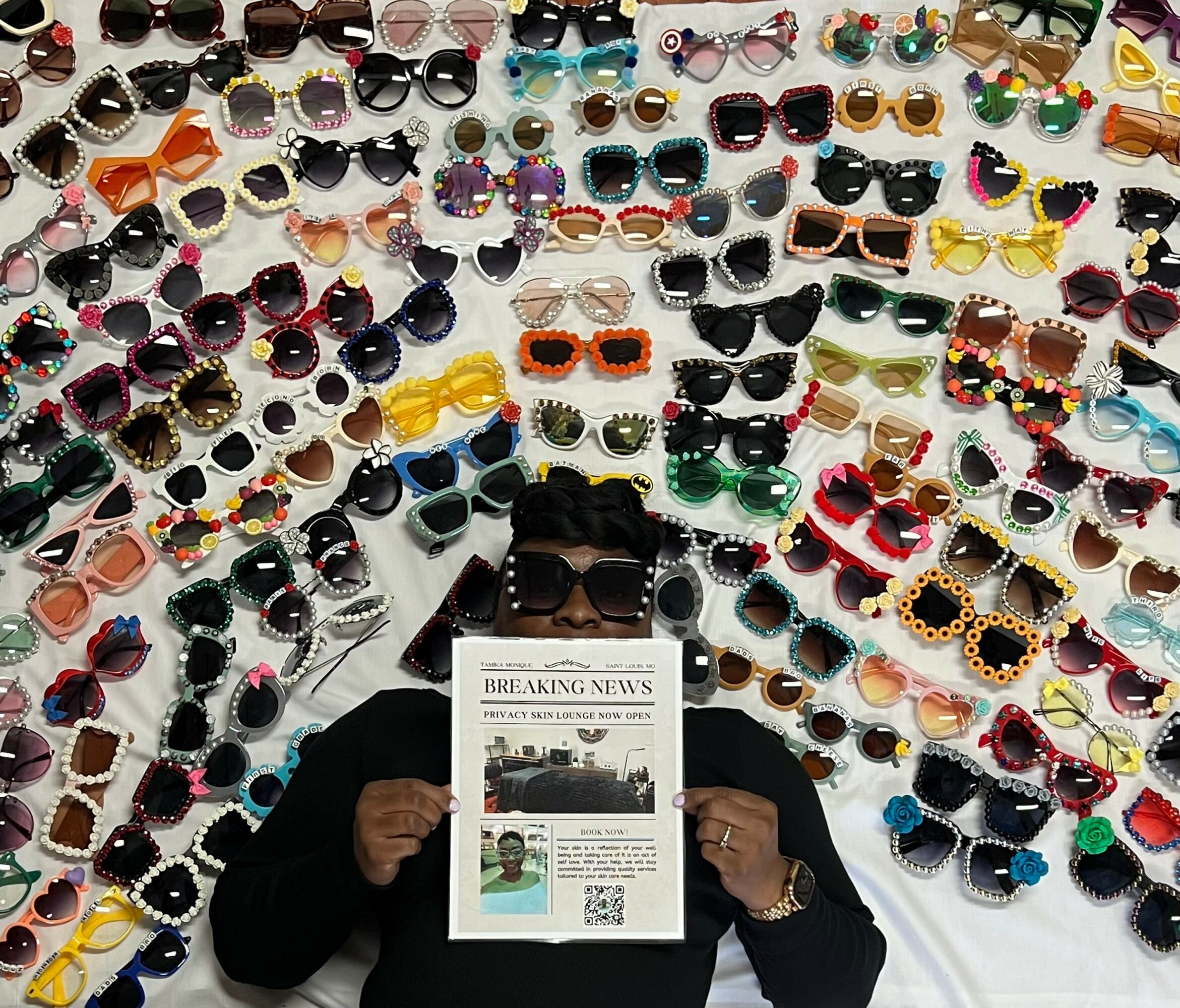 Person lying down holding a newspaper surrounded by colorful sunglasses arranged in a pattern.