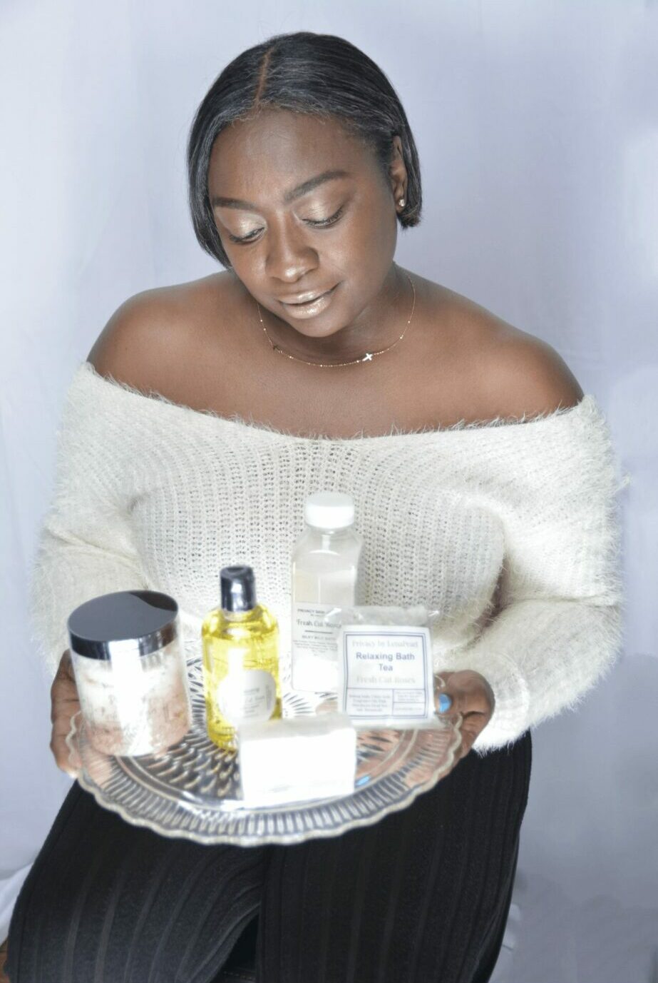 Young woman with short hair holding a tray of skincare products, wearing an off-shoulder sweater and black pants.