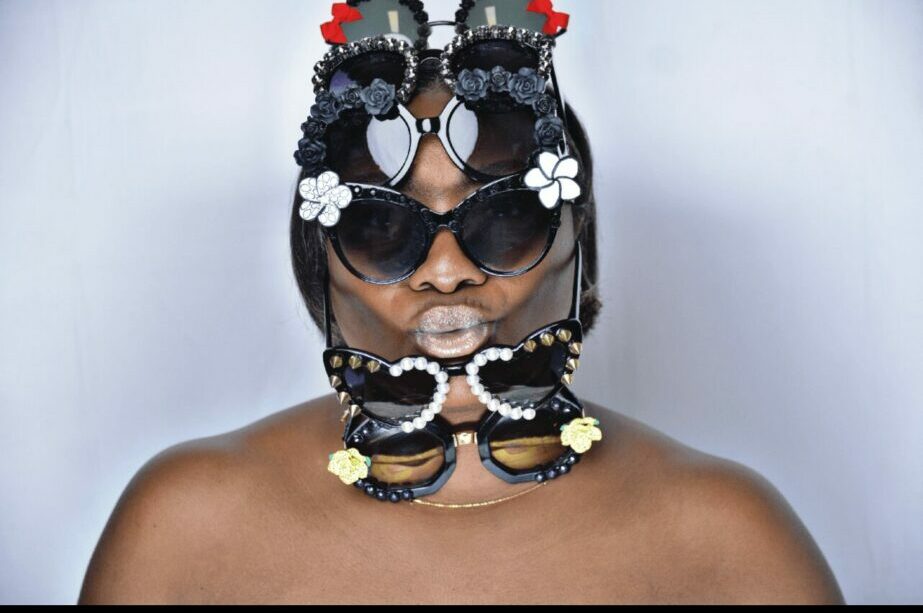 Person wearing large sunglasses and elaborate headpiece with flowers and beads, against a plain background.