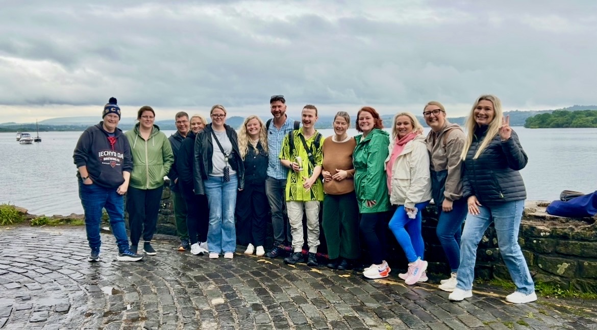 Group of fifteen people standing on a cobblestone path near a body of water with cloudy sky.