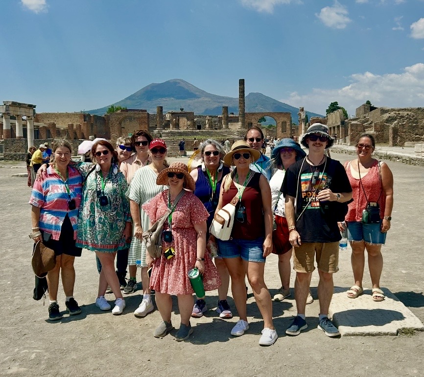 Group of tourists standing outdoors with ancient ruins and a mountain in the background, sunny day, some wearing hats and sunglasses.