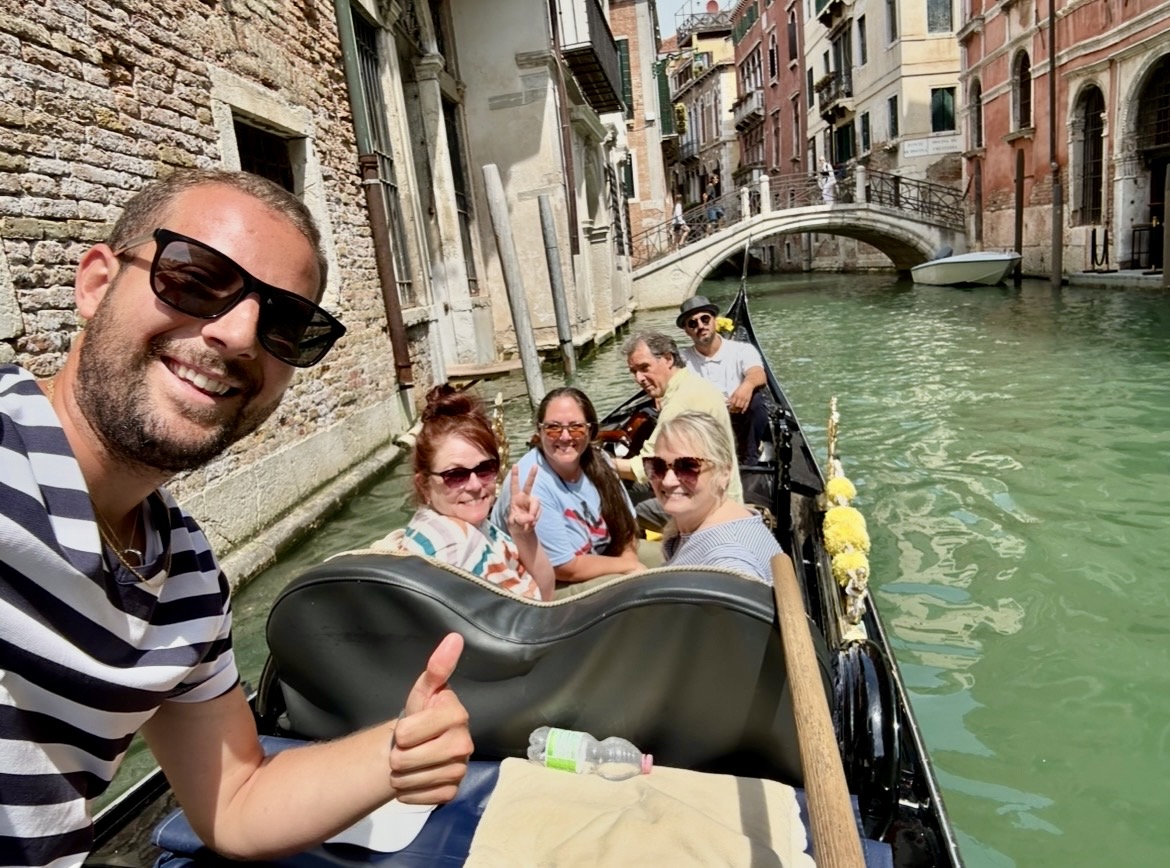 Group of people on a boat in a canal with buildings and a bridge in the background, smiling and enjoying the ride.