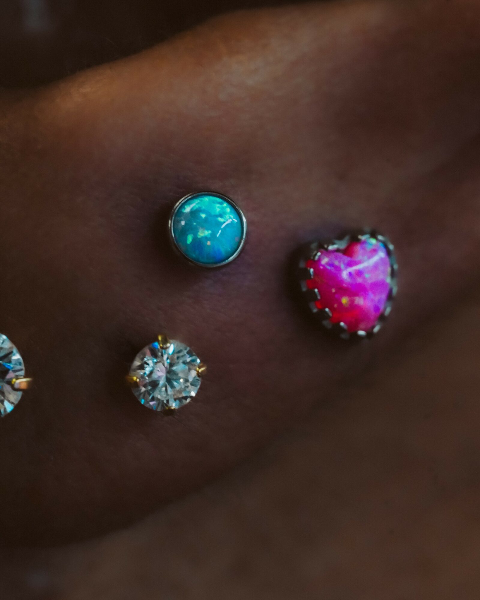 Close-up of skin with three colorful, decorative body piercings, including a blue, pink heart-shaped, and clear gem.