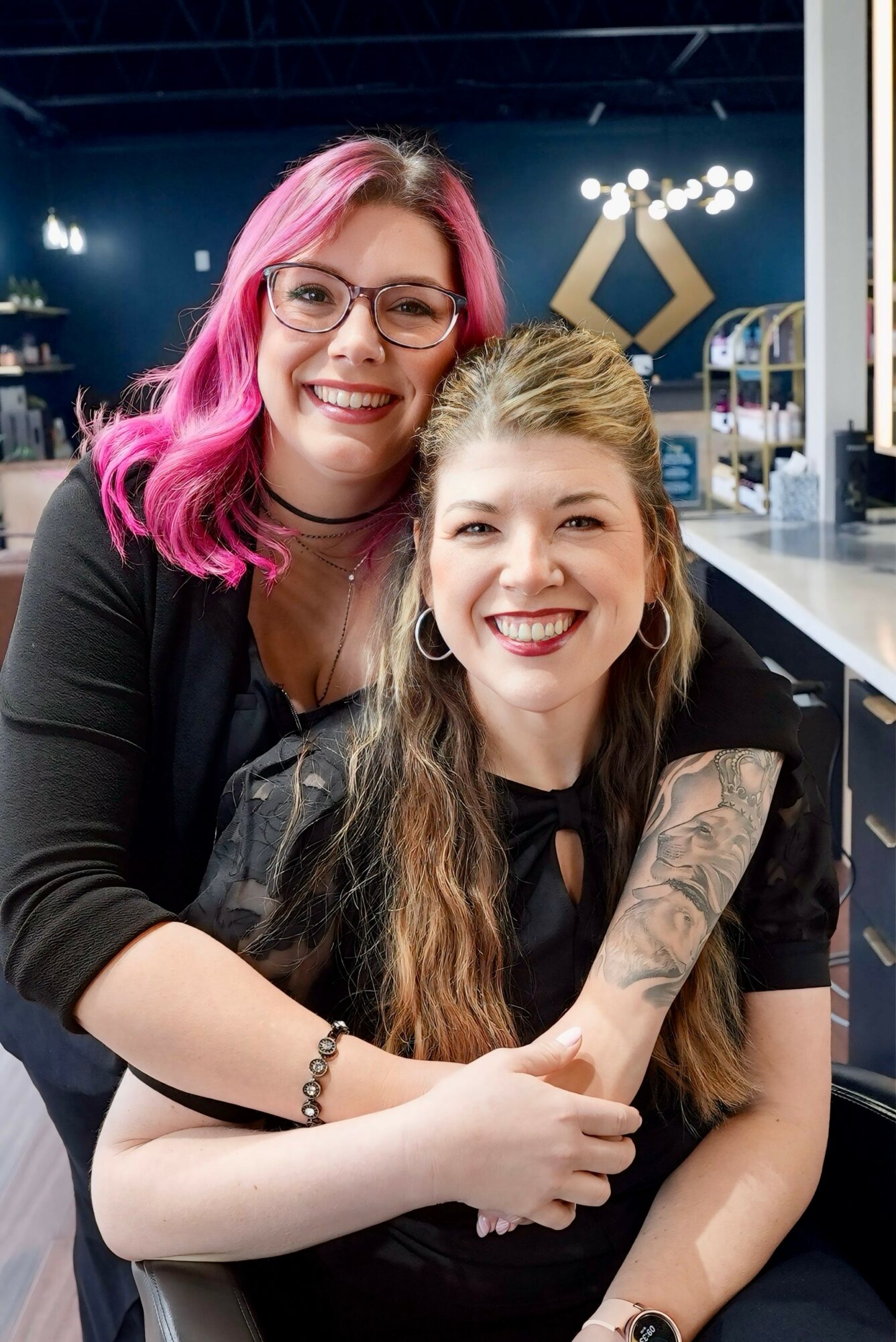 Two smiling women with long hair, one with pink hair and glasses, the other with long brown hair, in a modern indoor setting.