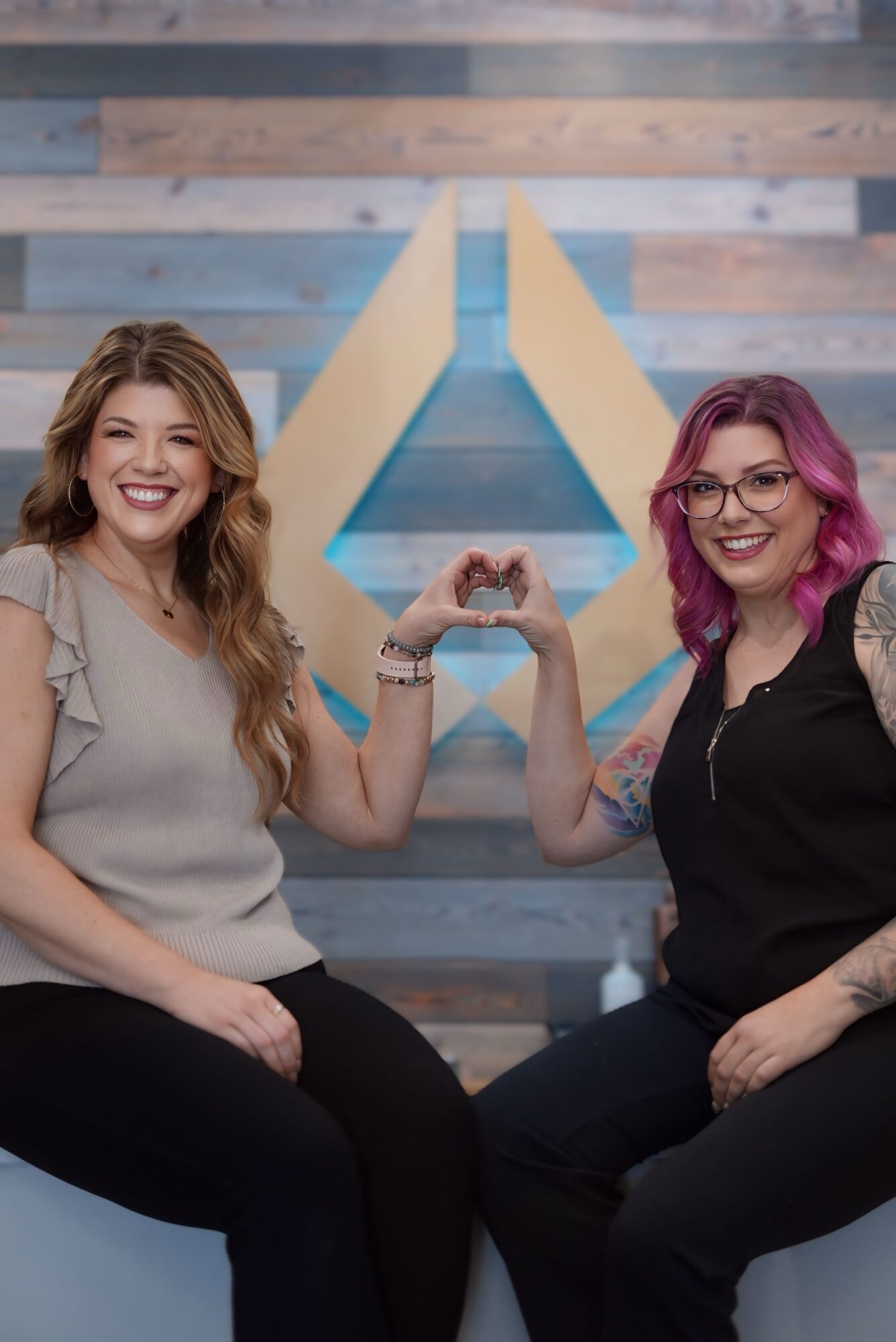 Two women sit side by side, smiling and holding hands with a heart shape between them, in front of a colorful background.
