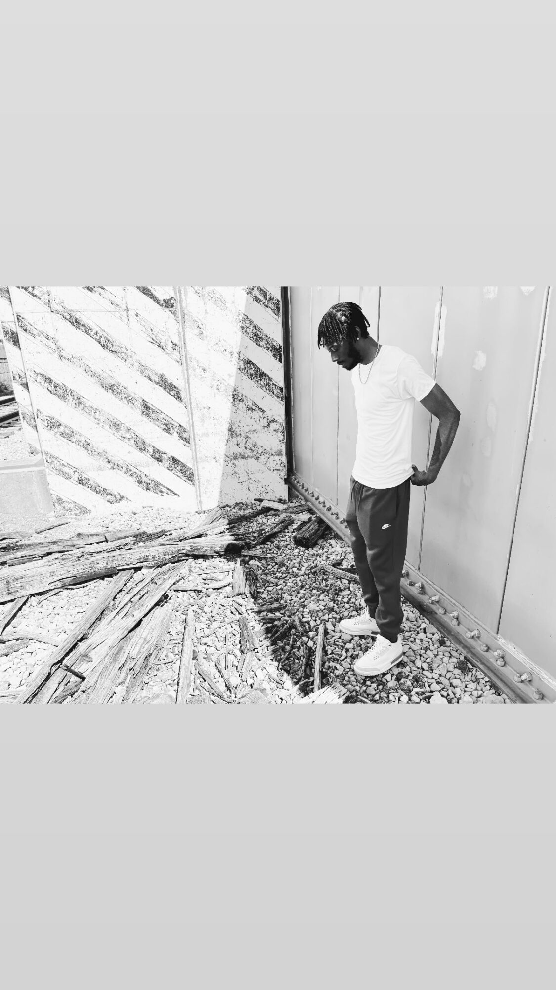 Person standing outdoors near a fence and wall, looking down at the ground, wearing a white shirt and dark pants.
