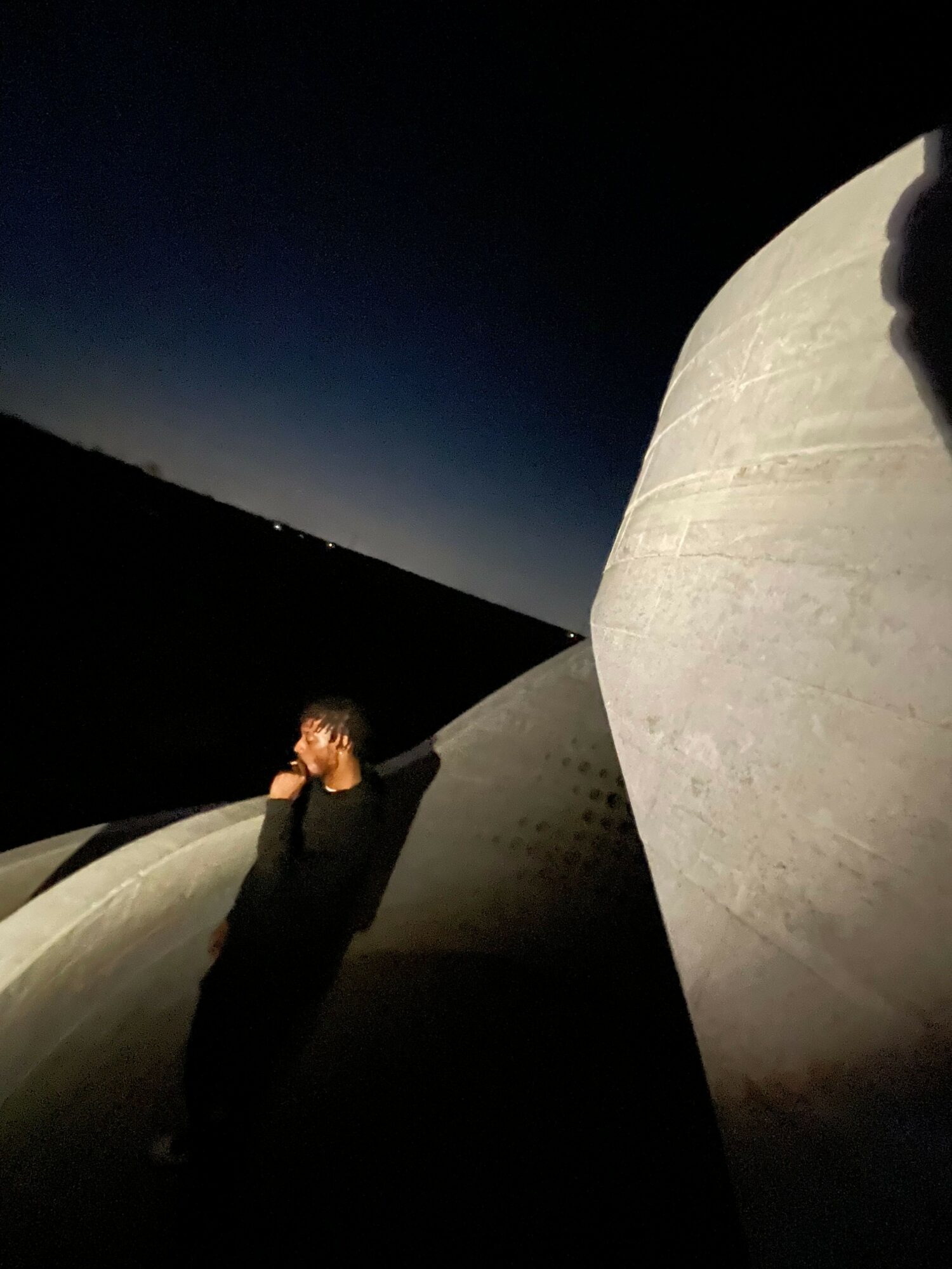 Person standing on a curved, concrete structure at night with a dark sky and a large spherical object above.