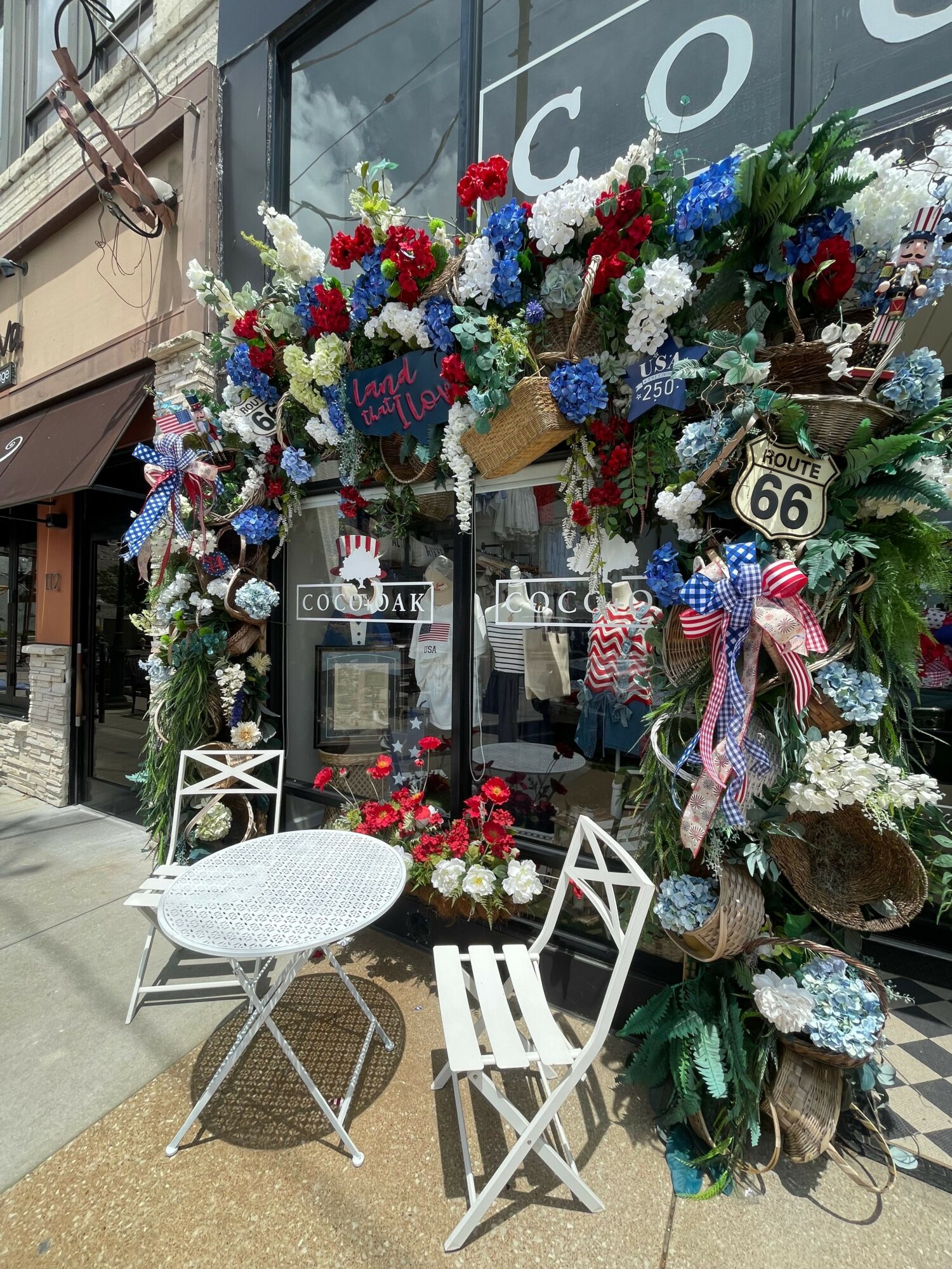 Decorative wreath with flowers, ribbons, and ornaments surrounds a window display with framed photos and a small table with chairs.
