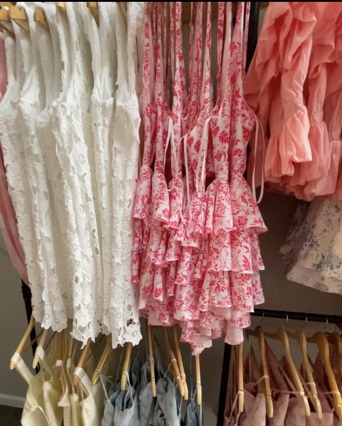Display of dresses with lace, floral, and ruffled designs hanging on a rack, in various colors including white, pink, and peach.