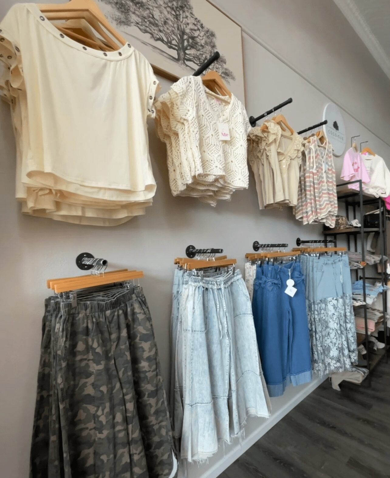 Clothing display with tops, skirts, and pants on hangers in a store, arranged on a wall and shelf.