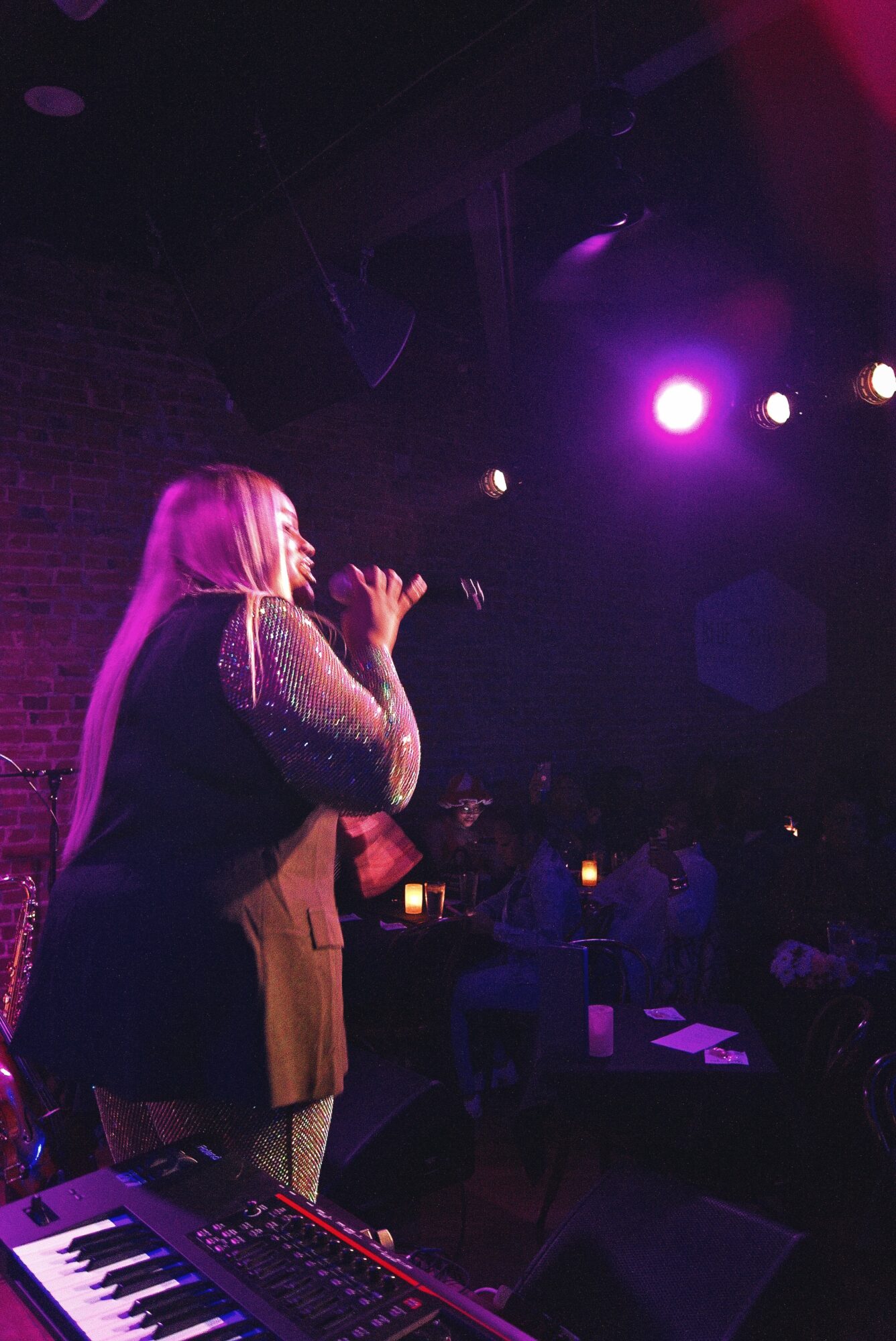 A woman with long hair sings into a microphone on stage, illuminated by purple and pink stage lights, with an audience in the background.