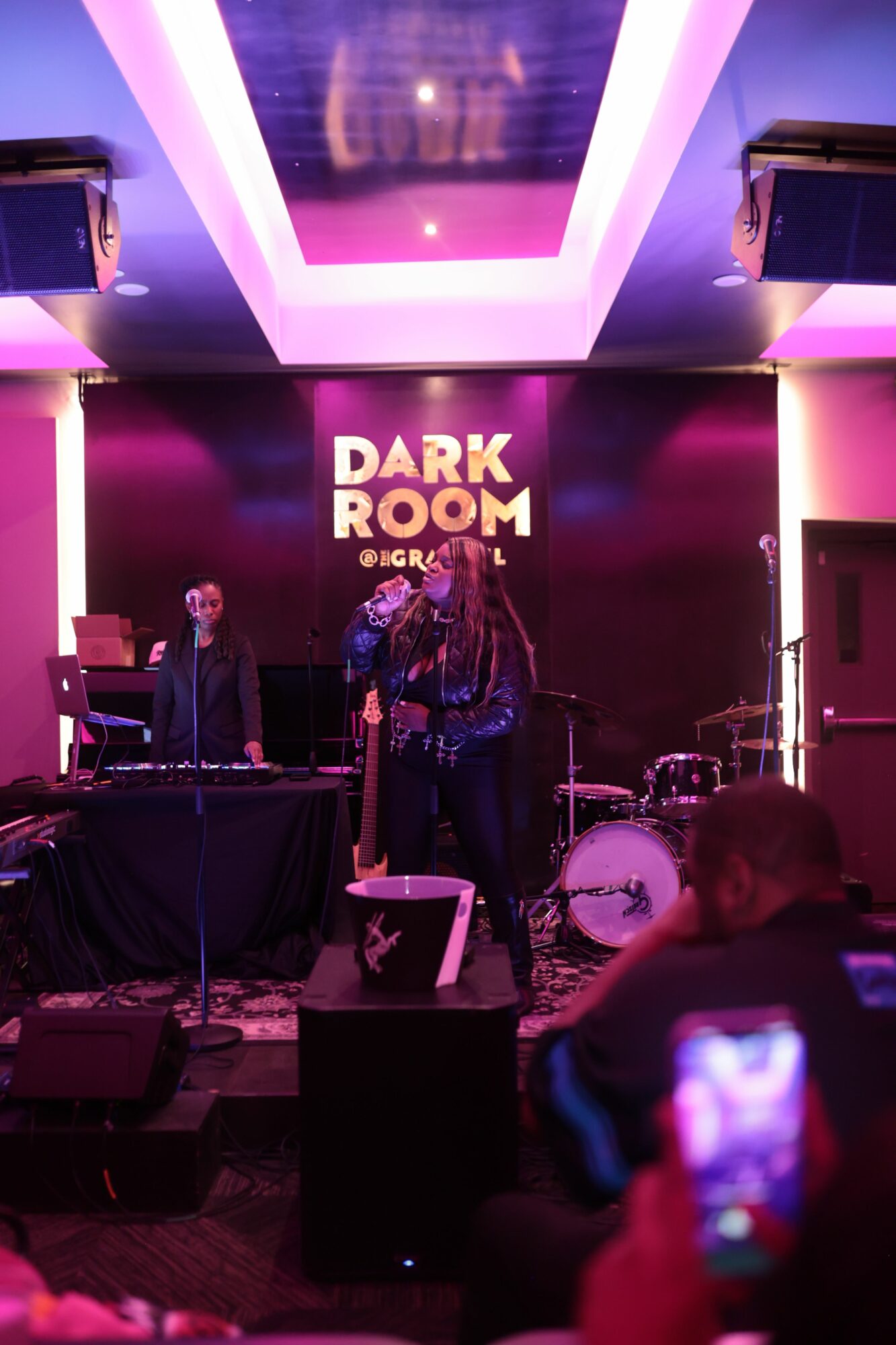 People on stage in a dark room with purple lighting, a sign reads 'DARK ROOM', audience in foreground, some taking photos.