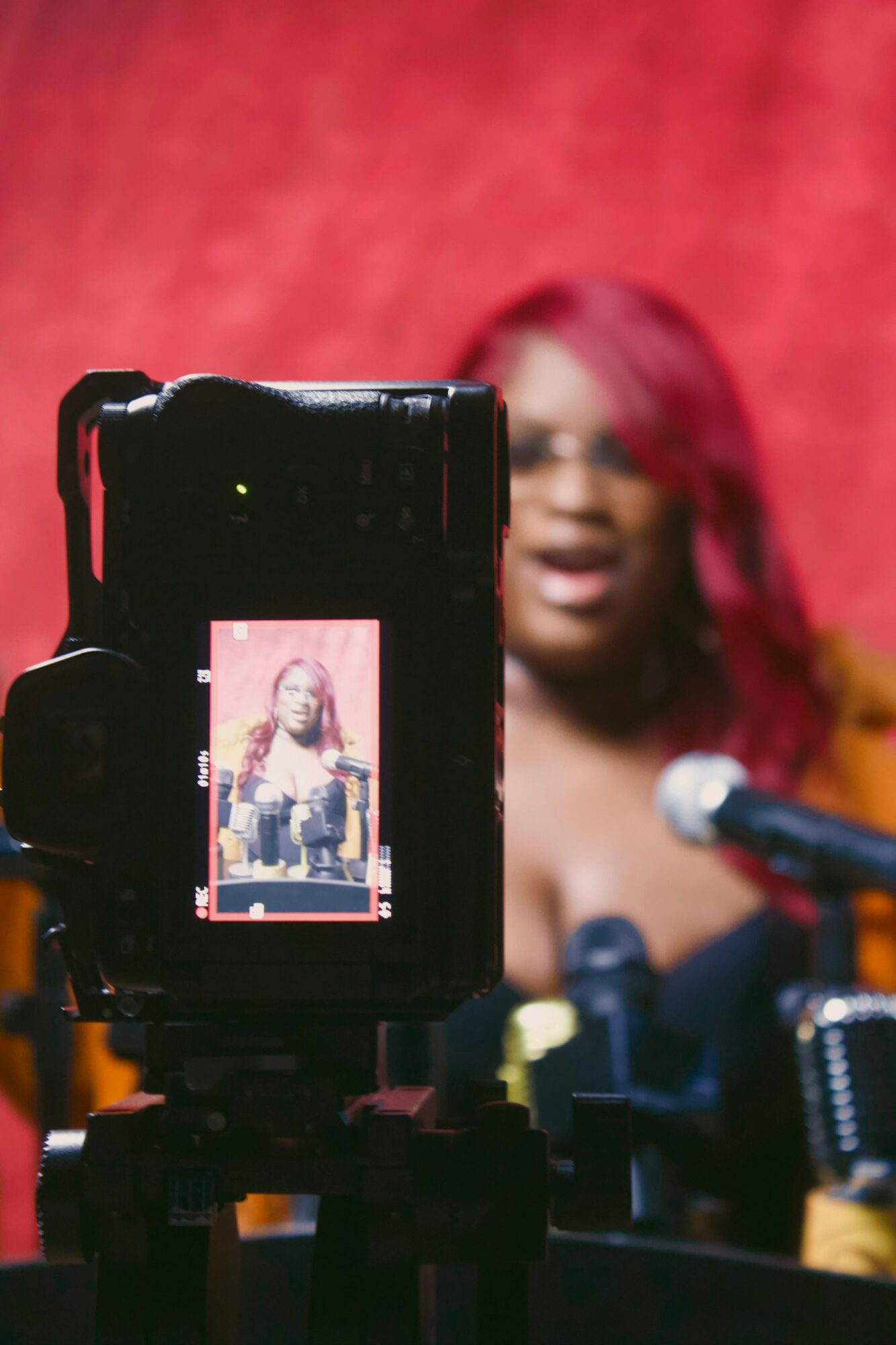 Person with red hair speaking into a microphone, seen through a camera screen, against a red background.