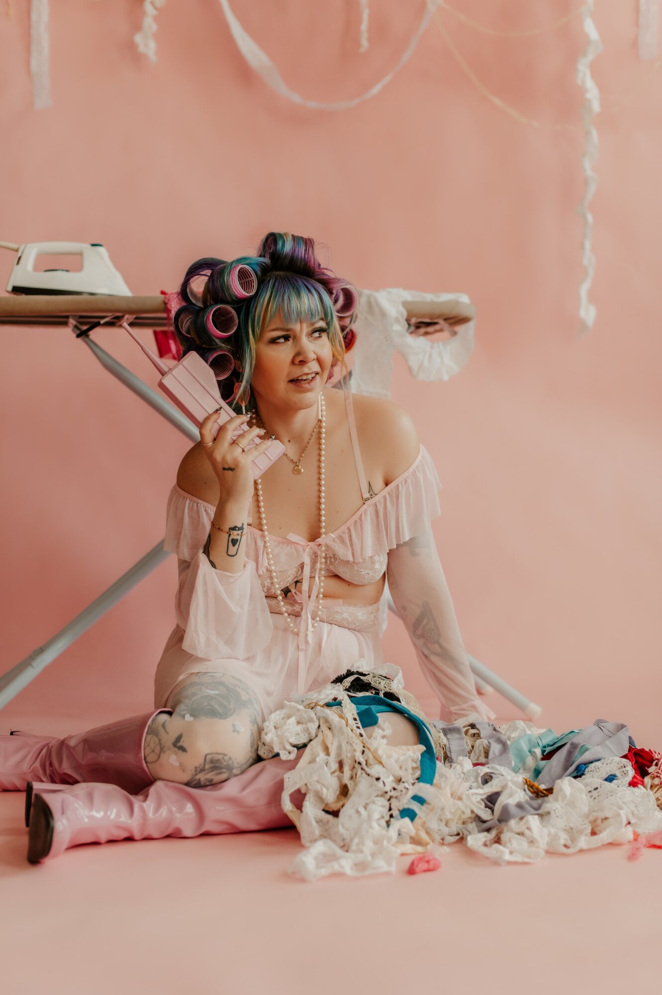 Young woman with colorful hair rollers, sitting on a pink surface, surrounded by clothes and accessories, against a pink background.