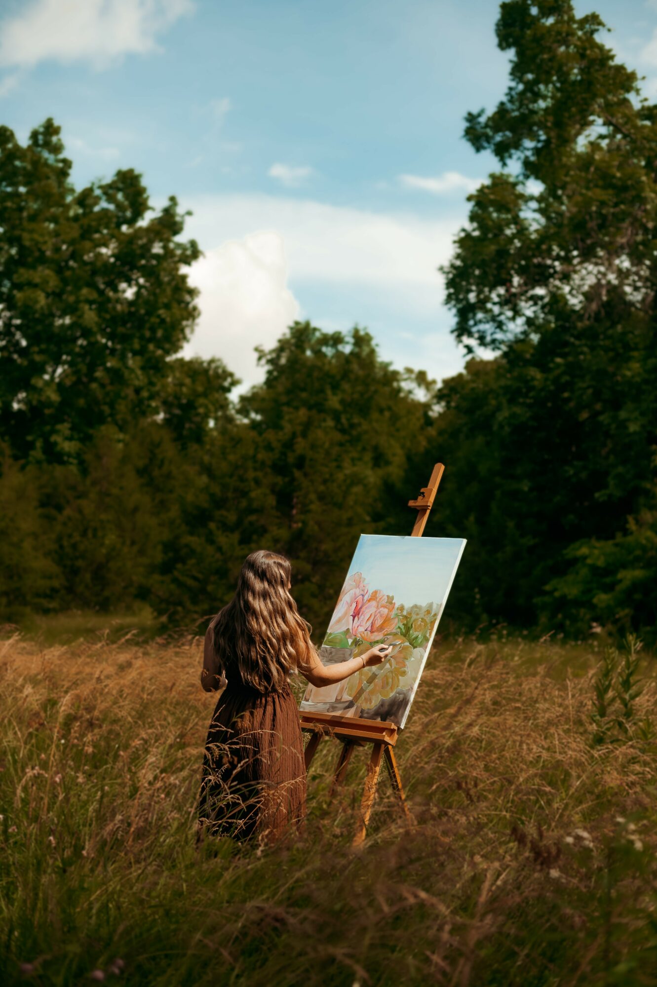 Girl painting outdoors in a grassy field with trees and blue sky in the background.