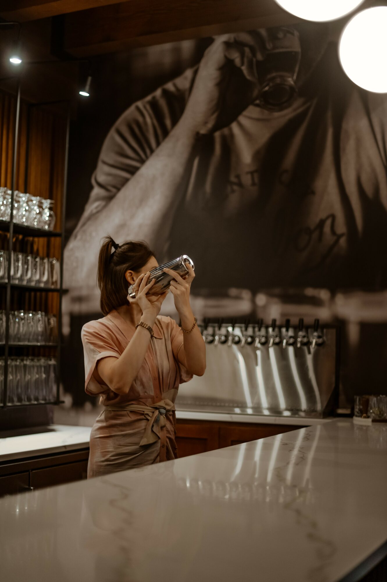 A woman in a pink shirt and apron drinks from a glass in a cafe with a large black-and-white photo of a person on the wall behind her.