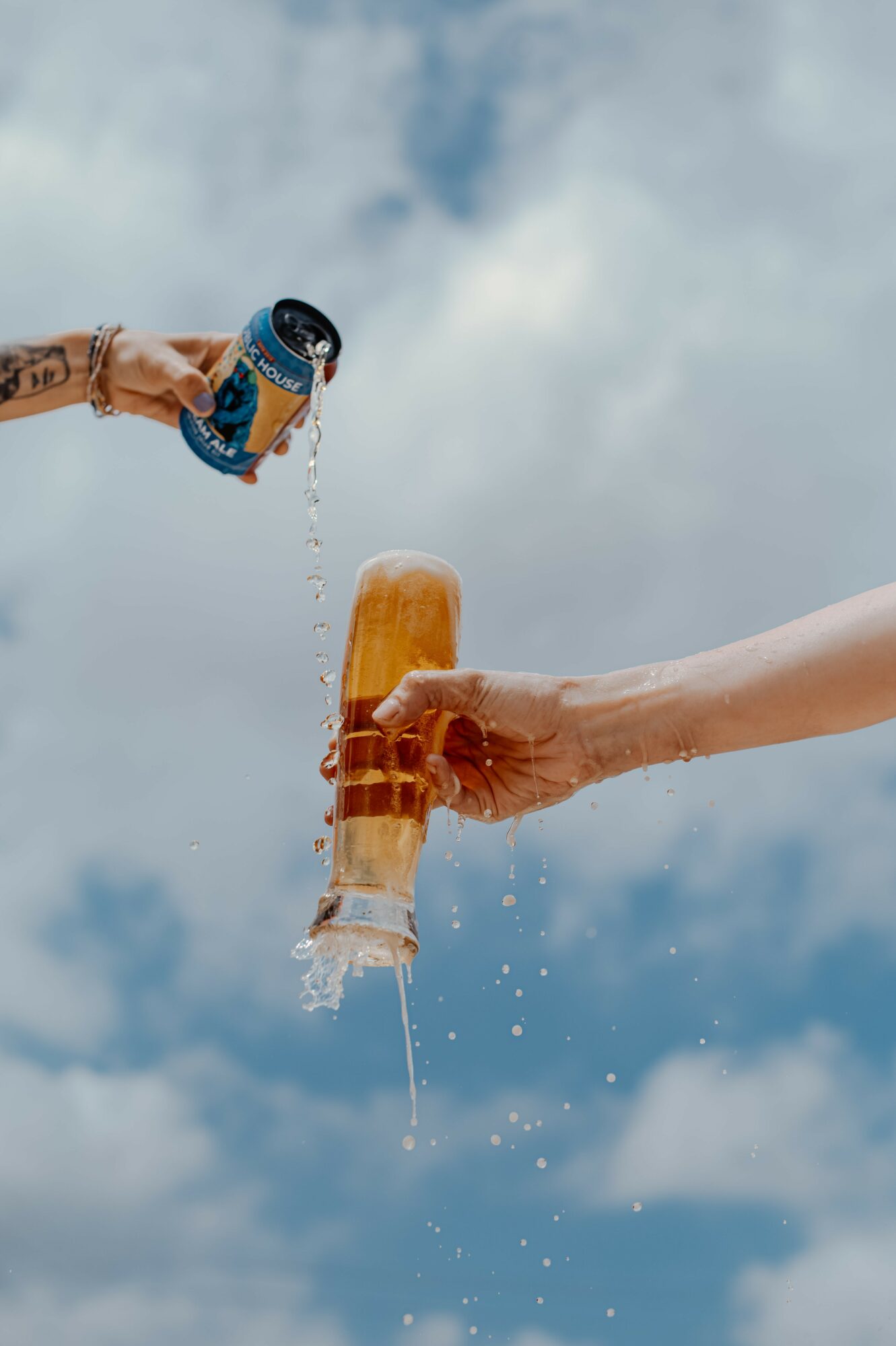 Two hands hold drinks, pouring liquid into each other against a cloudy sky background.