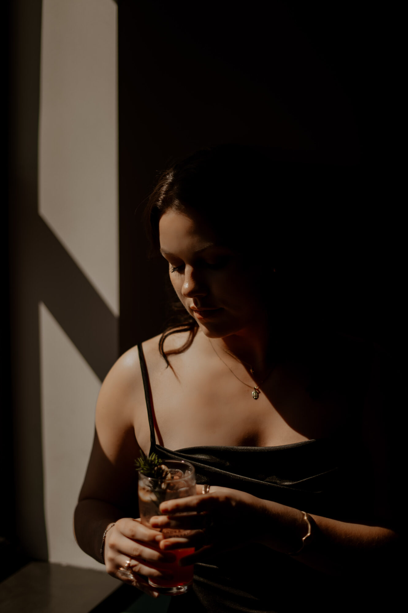 Woman holding a drink, illuminated by light from a window, with dark background and shadows.