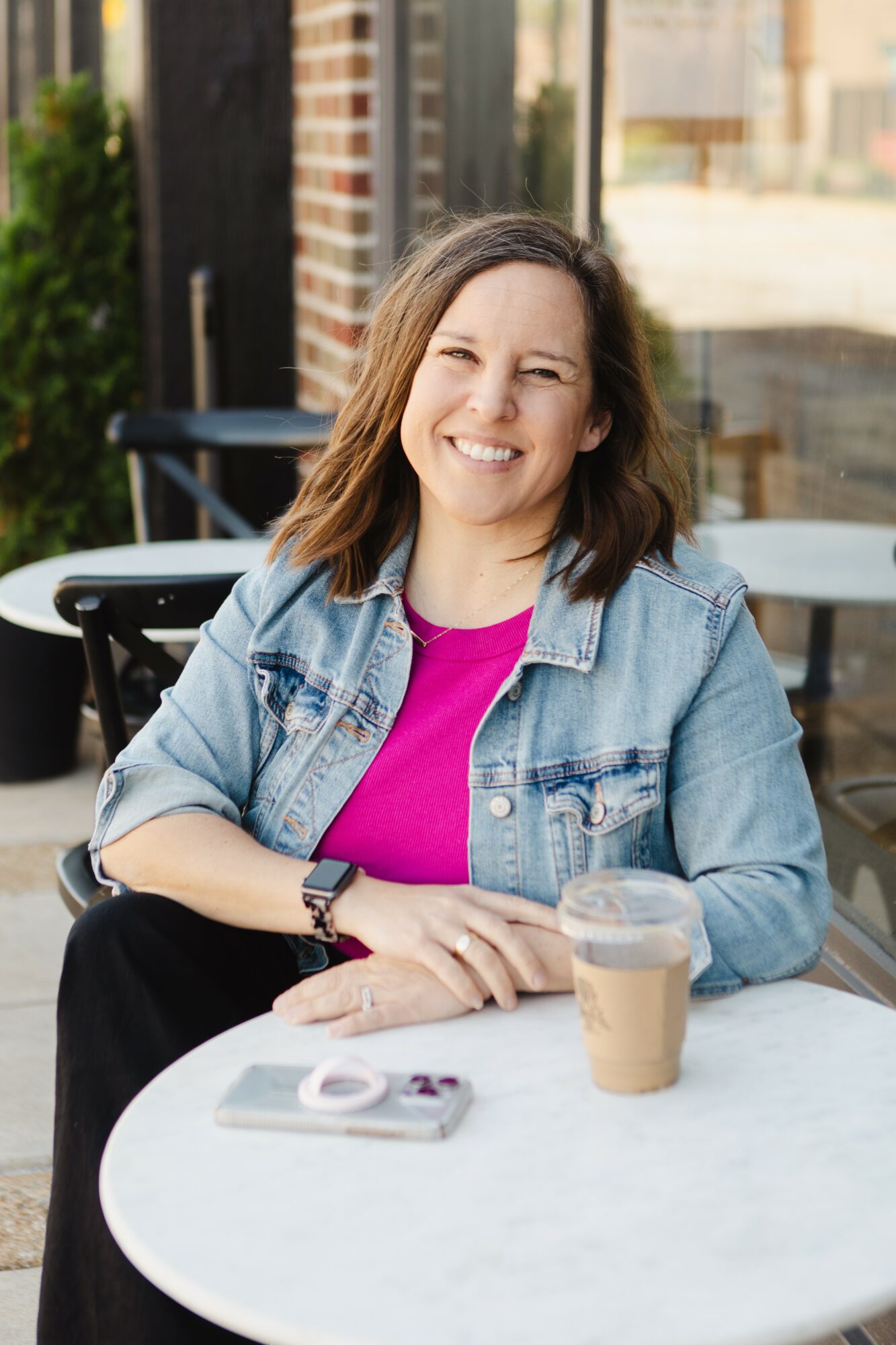 Woman with shoulder-length hair smiling, sitting at a table outdoors with a coffee drink and a camera, wearing a denim jacket and pink top.