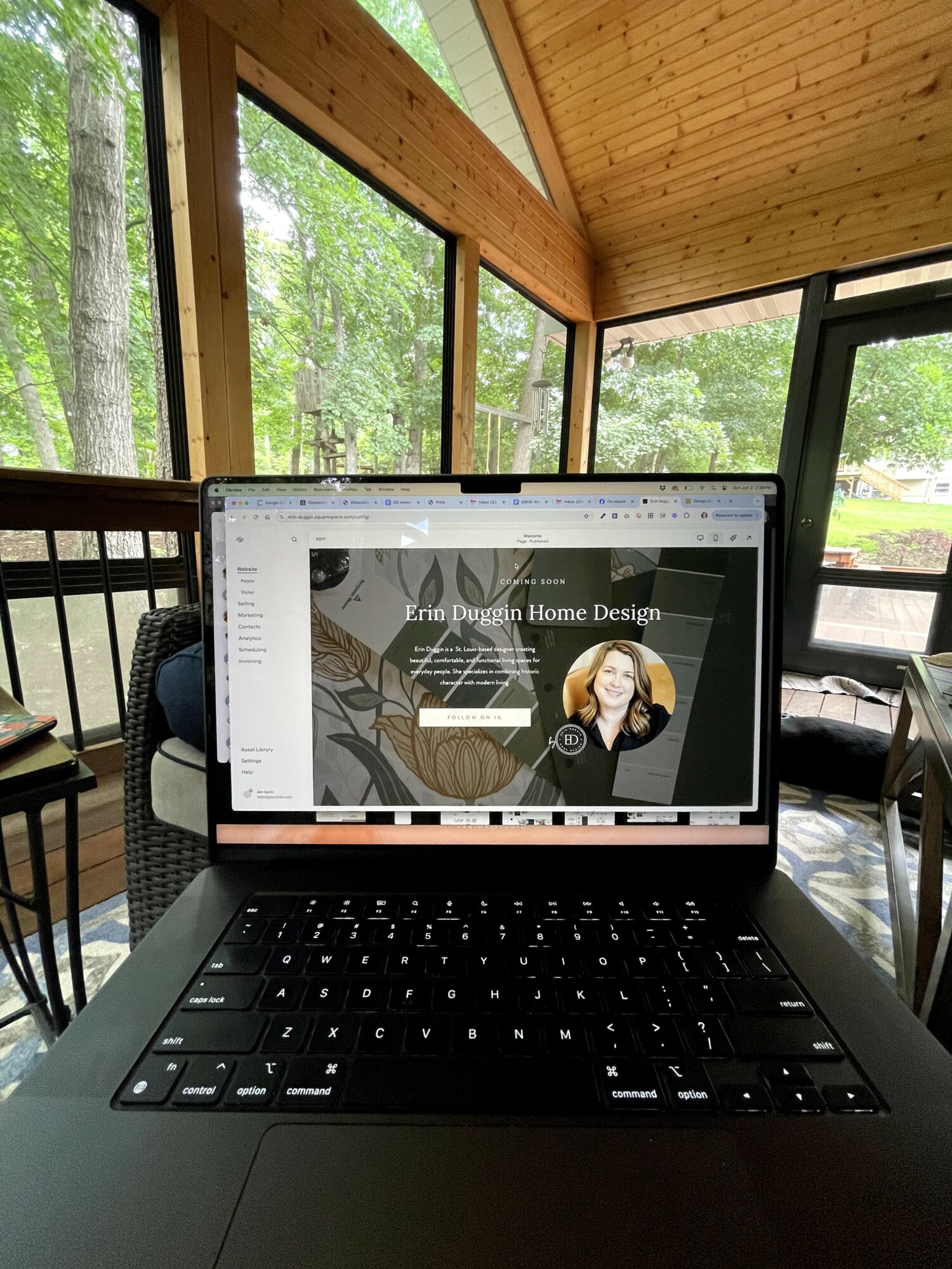 Laptop on table displaying a home design website with a woman’s photo, in a sunlit room with large windows and wooden ceiling.