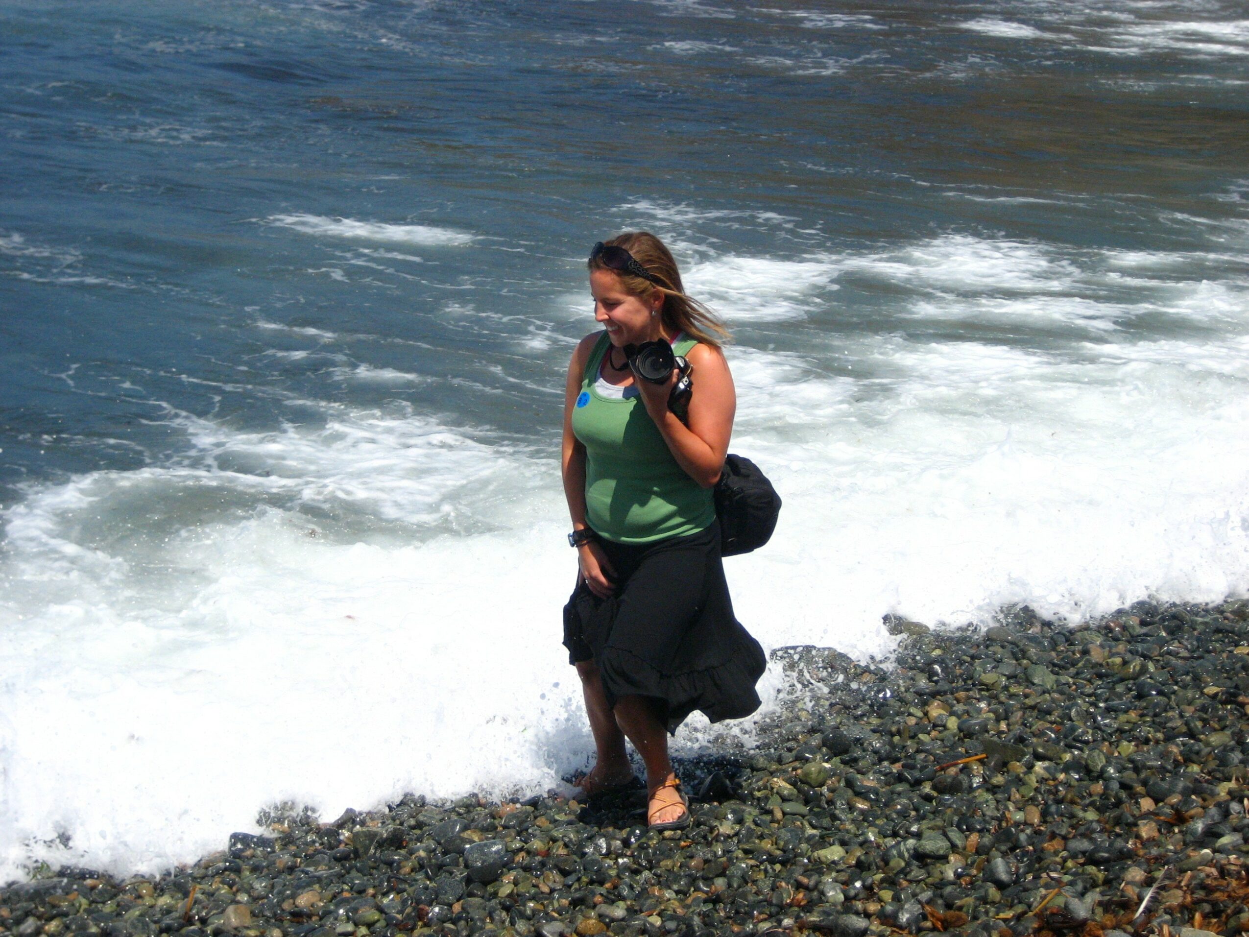 Woman walking on rocky beach near water, wearing a green top, black skirt, and sunglasses, carrying a black bag.