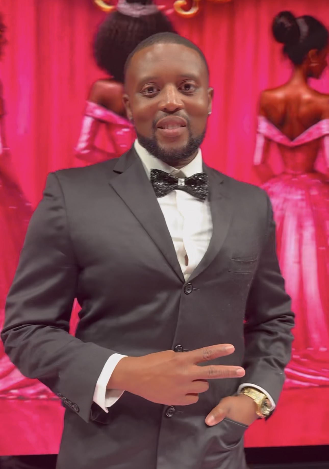 Man in a black suit and bow tie making a peace sign, standing in front of a pink curtain with women in pink dresses in the background.