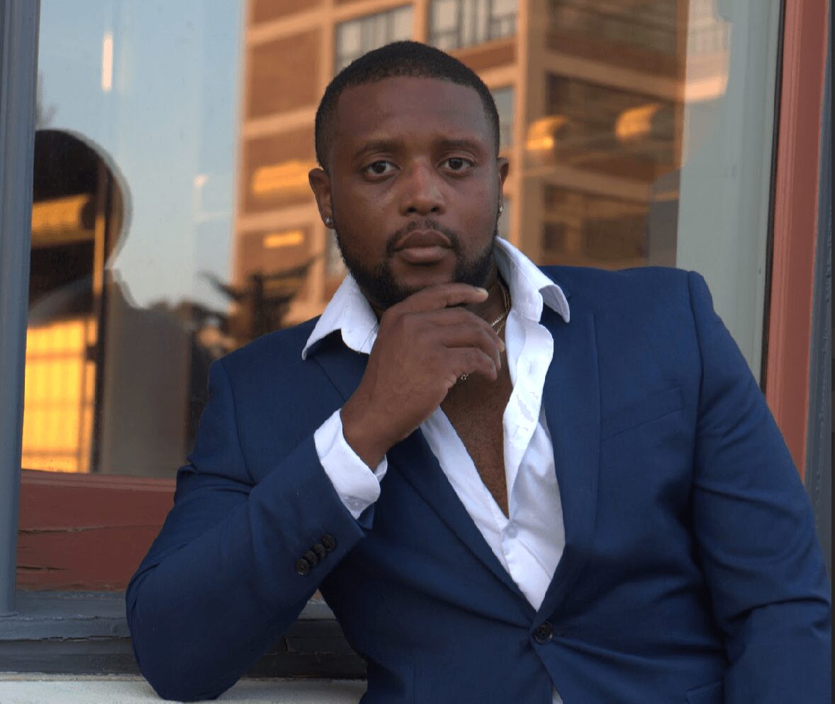 Man in a navy suit with white shirt, sitting outdoors, with buildings reflected in glass behind him.