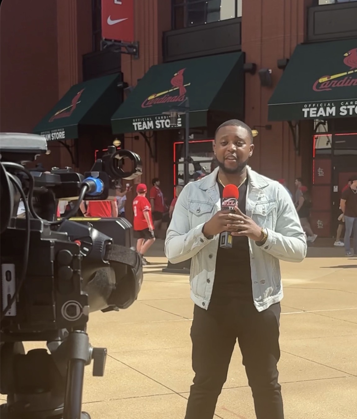 Man holding a microphone in front of a camera outside a store with green awnings and people walking by.