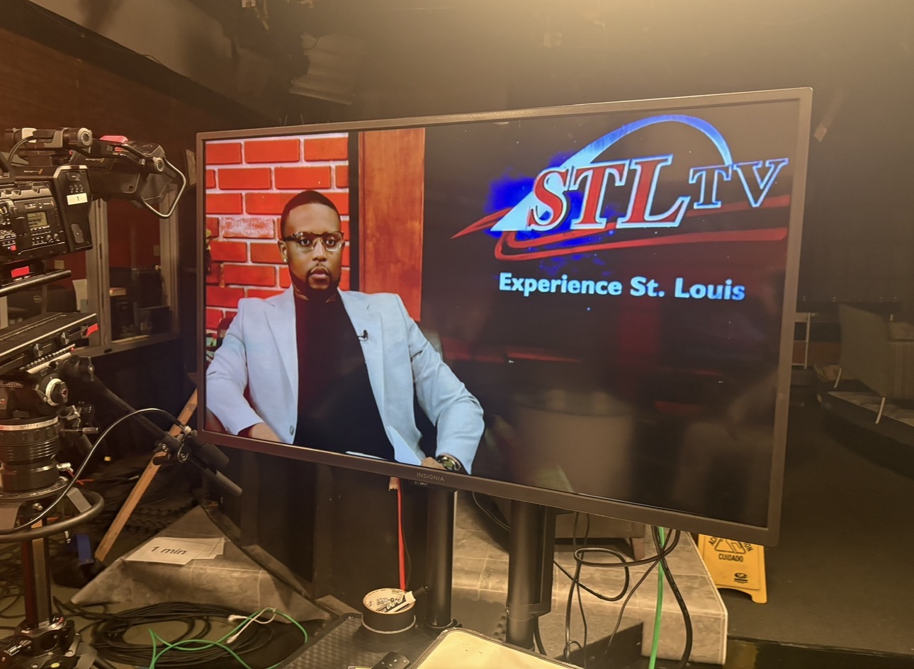 Person with glasses and a light jacket sitting in a studio with a large screen displaying 'STL TV' and 'Experience St. Louis'.
