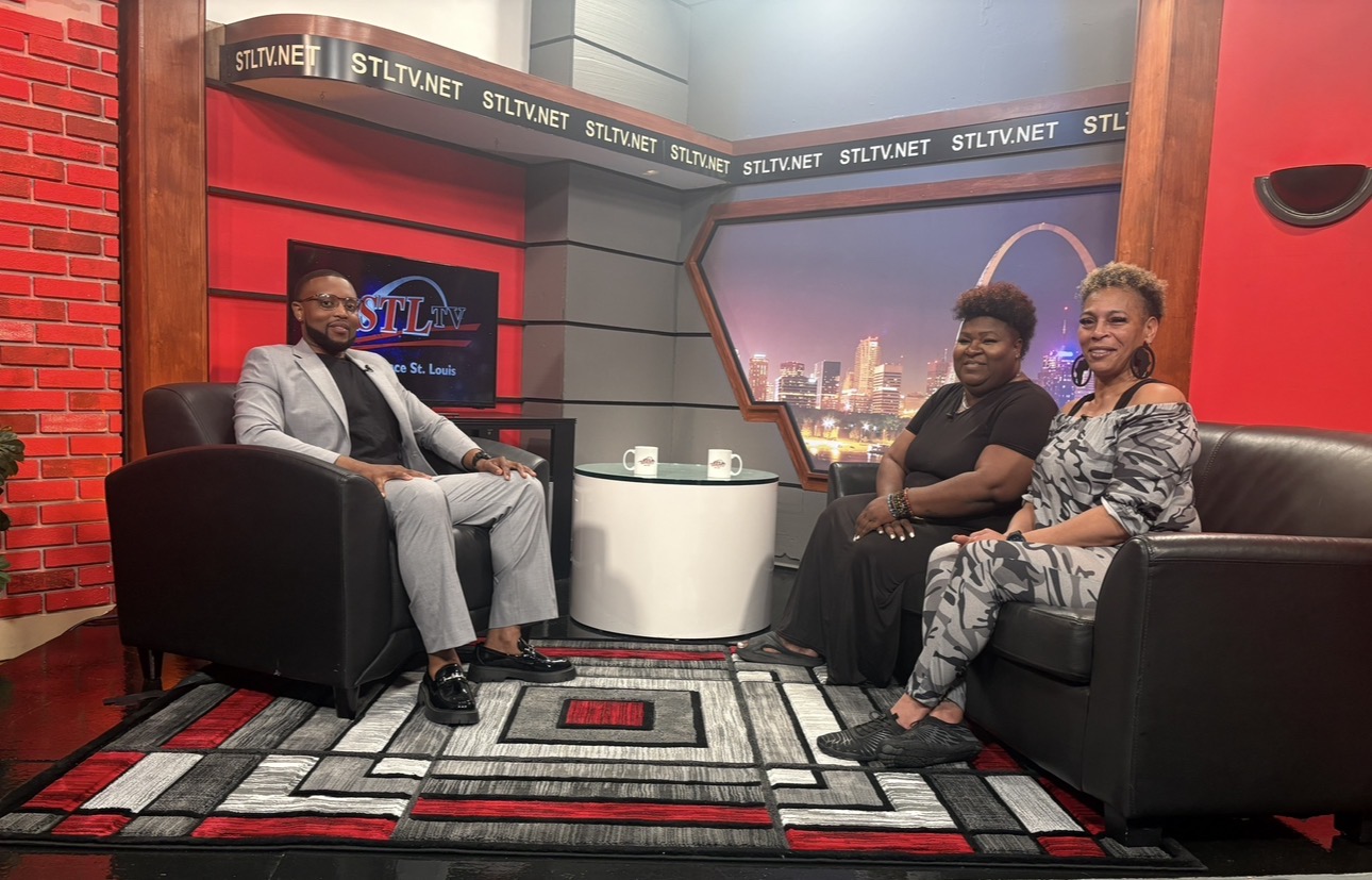 Three people seated on black sofas in a TV studio with a cityscape backdrop and red walls.