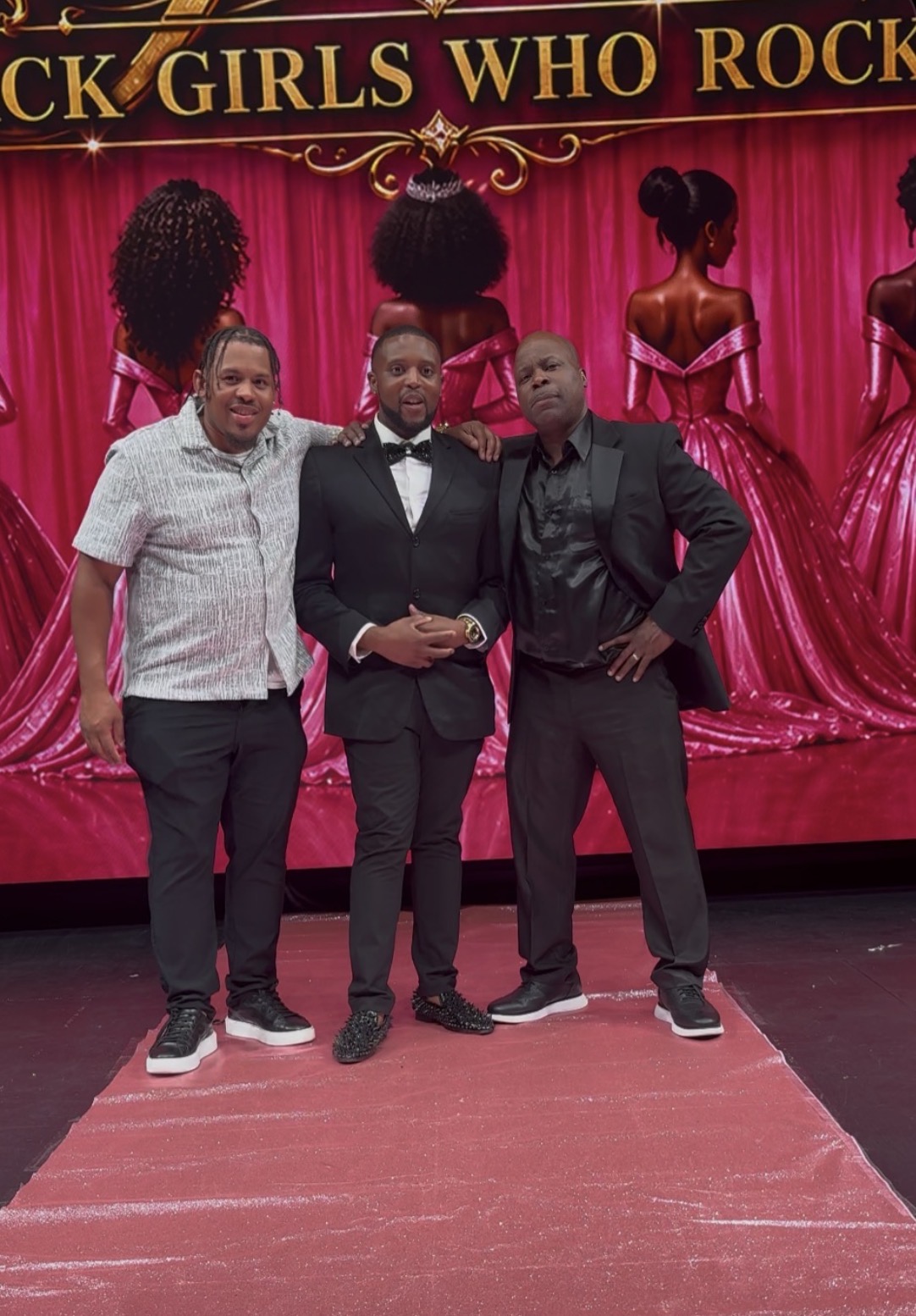 Three men stand on a stage with a red curtain and mannequins dressed in red gowns behind them.
