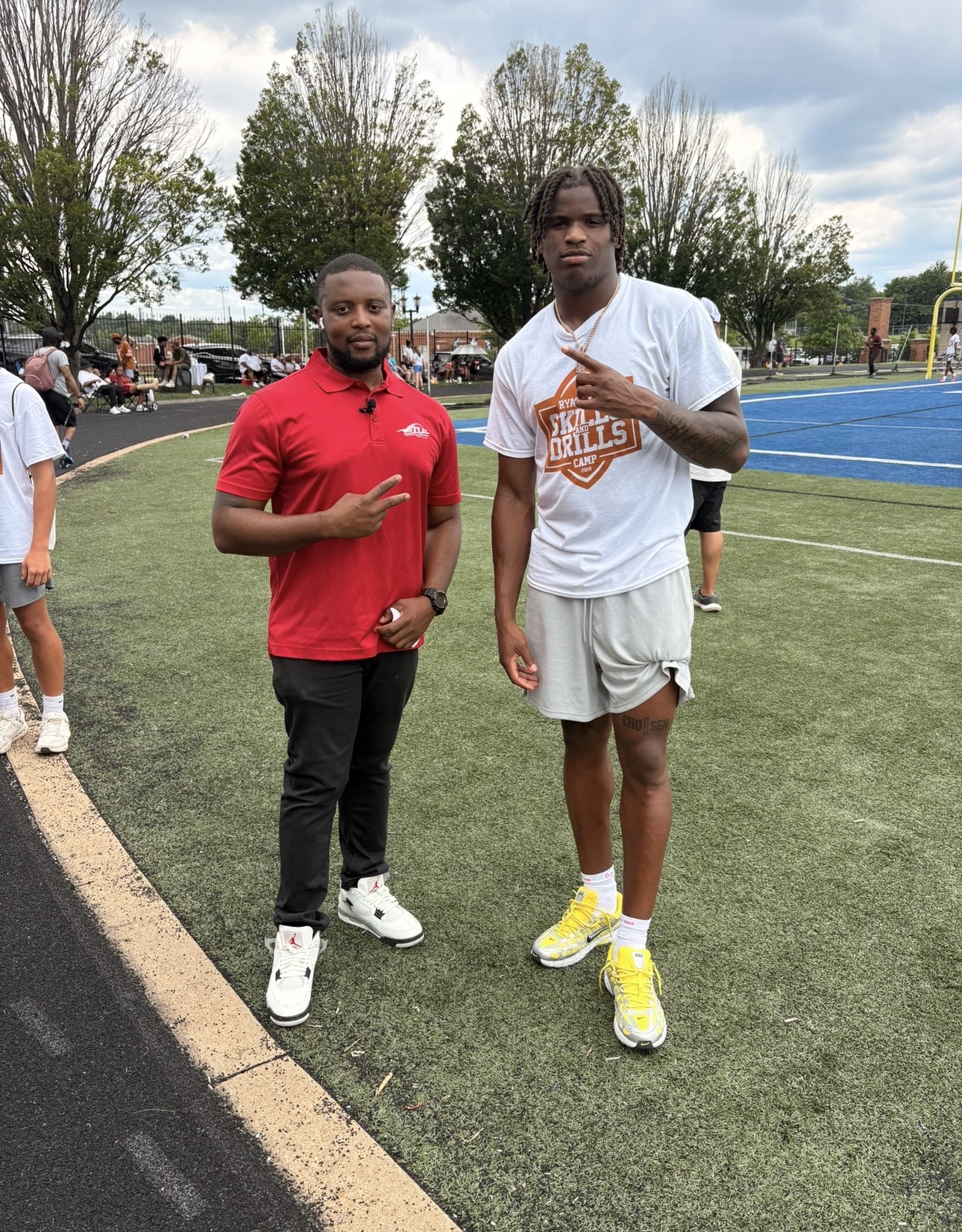 Two young men standing on a sports field, one in a red shirt and the other in a white shirt and shorts, with trees and a blue track in the background.