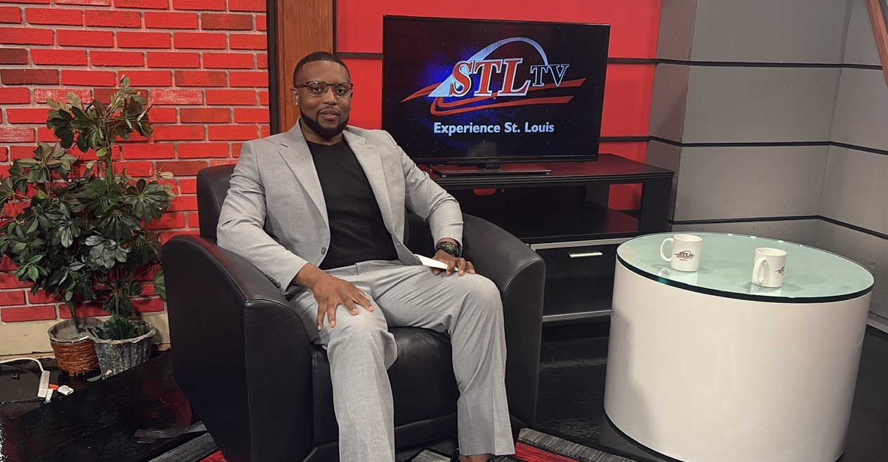 Man in light gray suit sitting on black armchair in studio with TV screen and white table nearby
