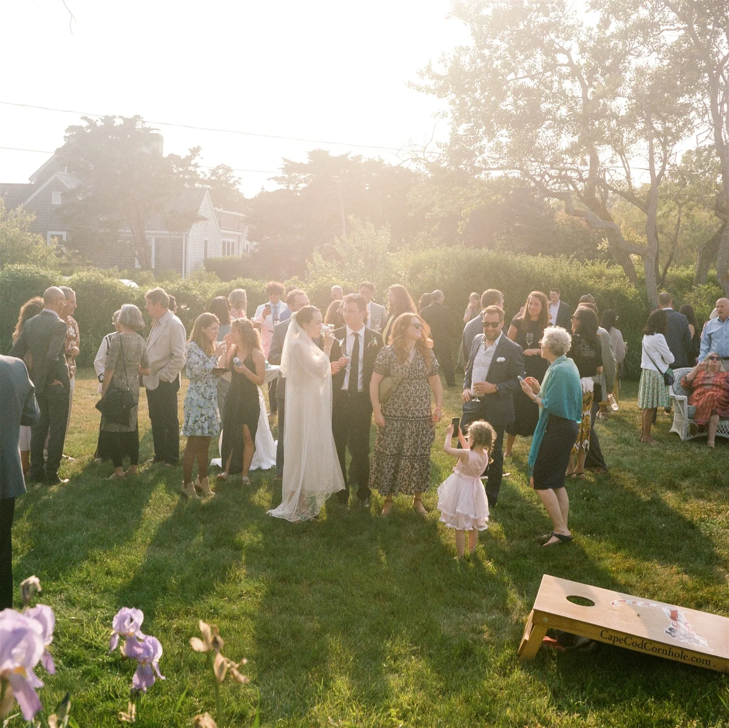 Group of people gathered outdoors on grass, some children, in sunlight with trees and houses in background.