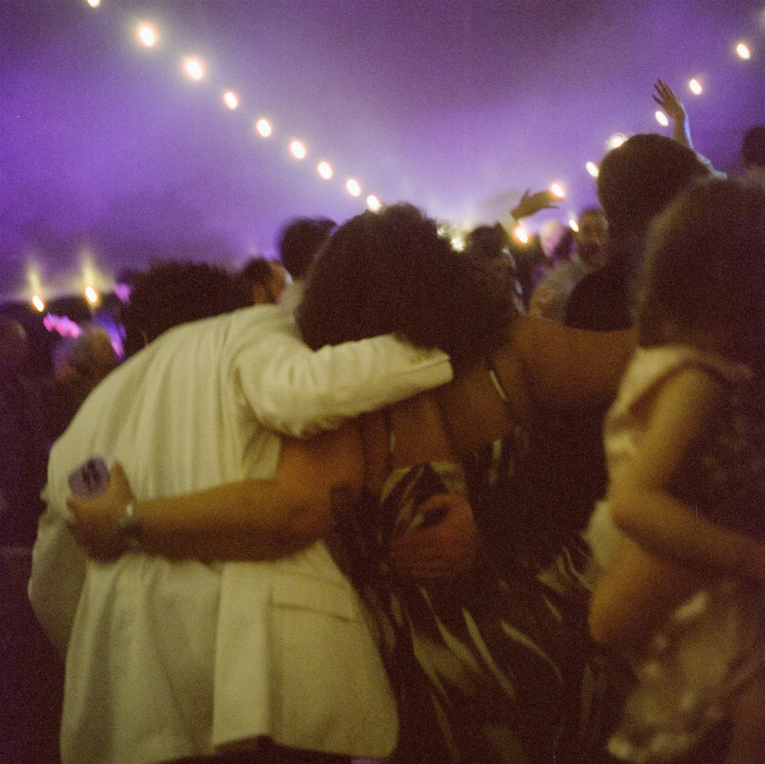 People at a concert or event, with one person hugging another, surrounded by string lights and a foggy atmosphere.
