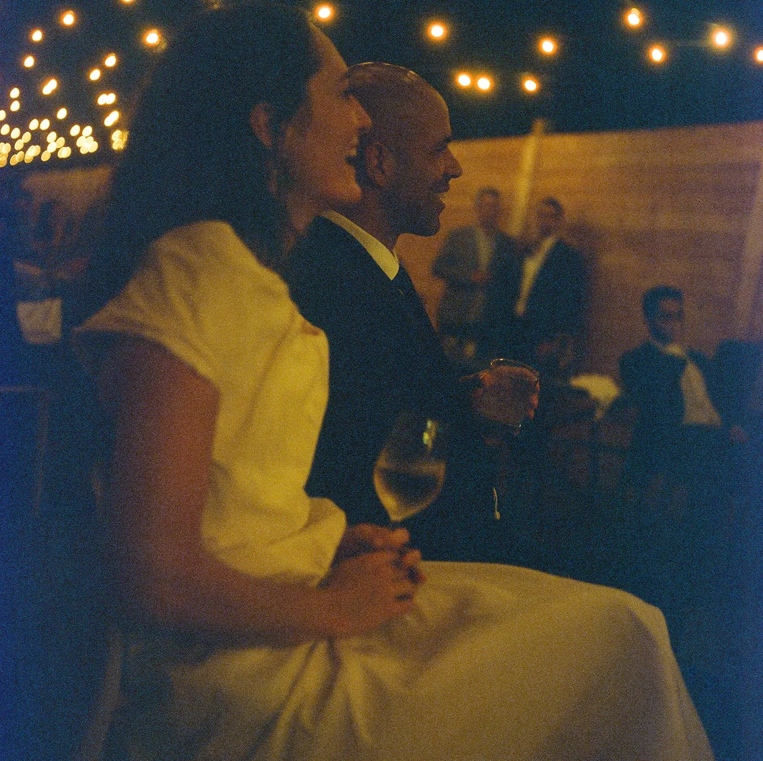 Two people sitting close together at an outdoor event, holding wine glasses, with string lights in the background.